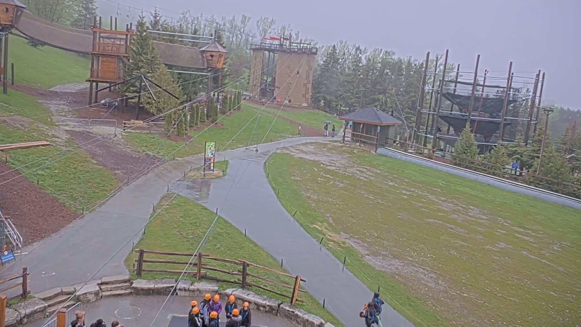 An outdoor adventure park with various climbing structures and zip lines is shown on a rainy day.