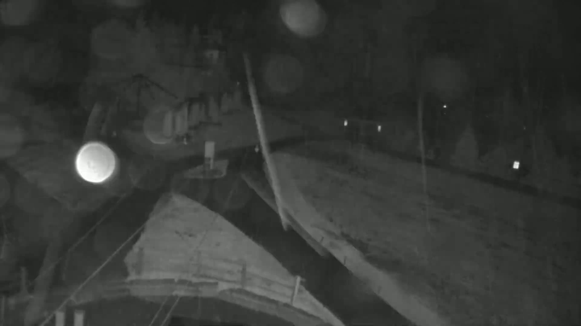 A nighttime, rainy view shows a dark, possibly snowy, ground with blurry lights in the distance and a dark object in the foreground.