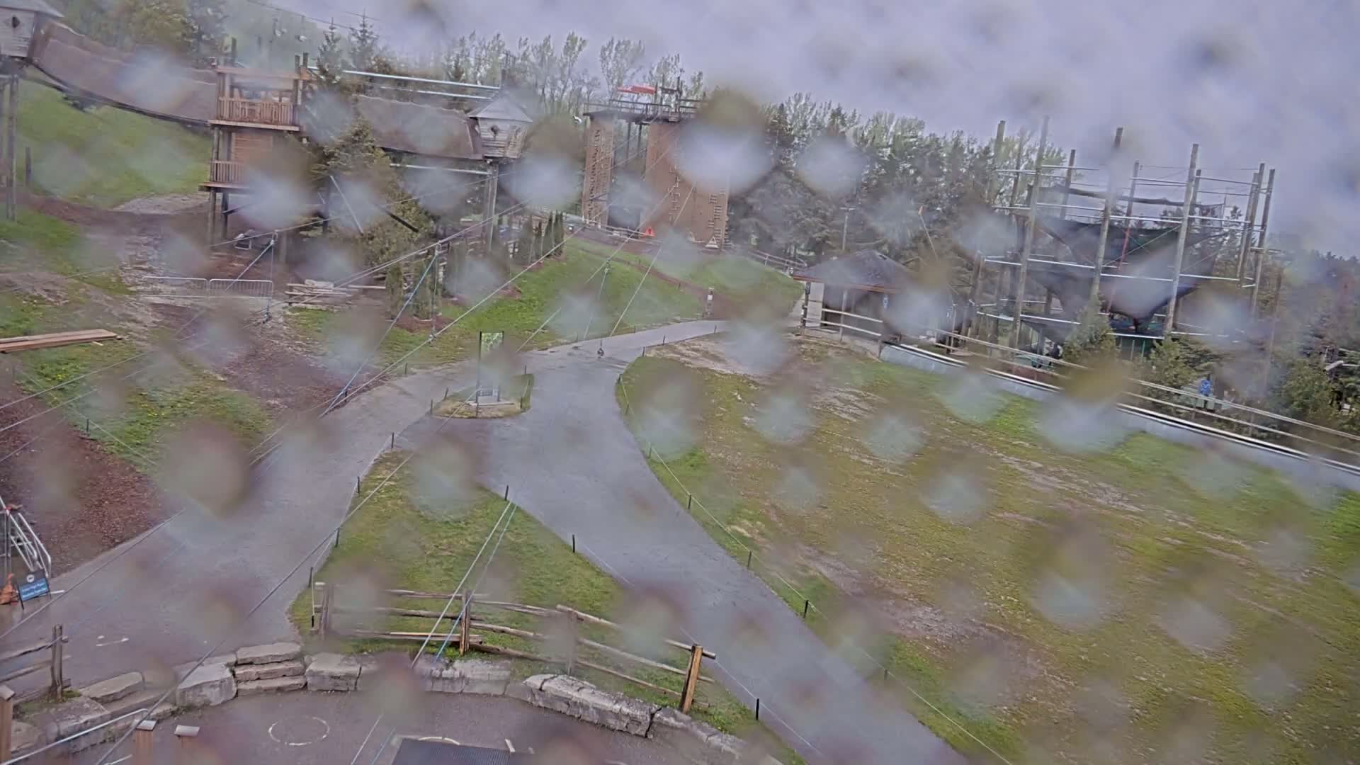 A rainy view of an outdoor adventure park with wooden structures, climbing walls, and zip lines, seen through a rain-covered window.