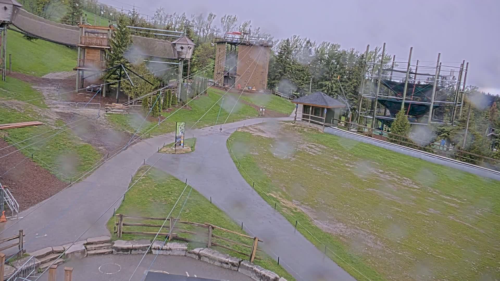 A rain-soaked outdoor adventure park features various climbing structures, zip lines, and paved pathways.