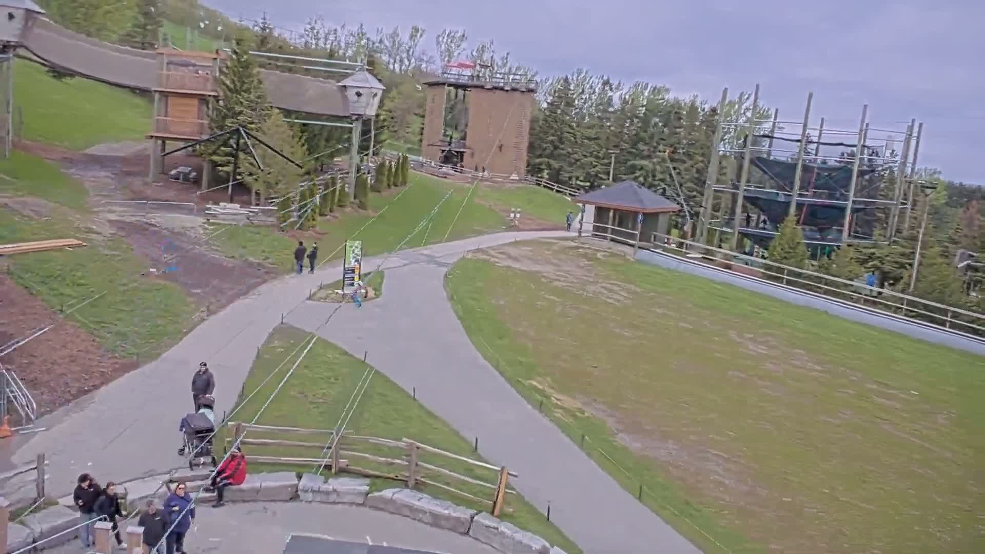 An outdoor aerial view shows a paved path winding through a grassy area with several people walking near various structures including a climbing tower and ropes course, under an overcast sky.