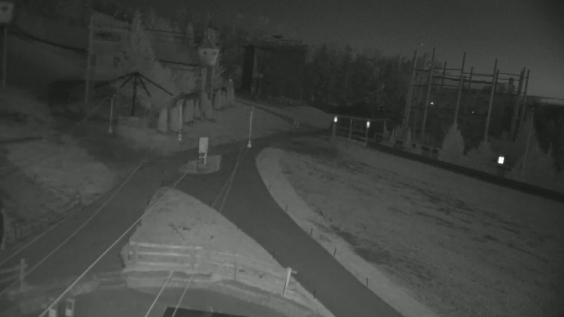 A nighttime, low-light image shows a paved road curving through an area with some structures and trees, under dark, clear skies.