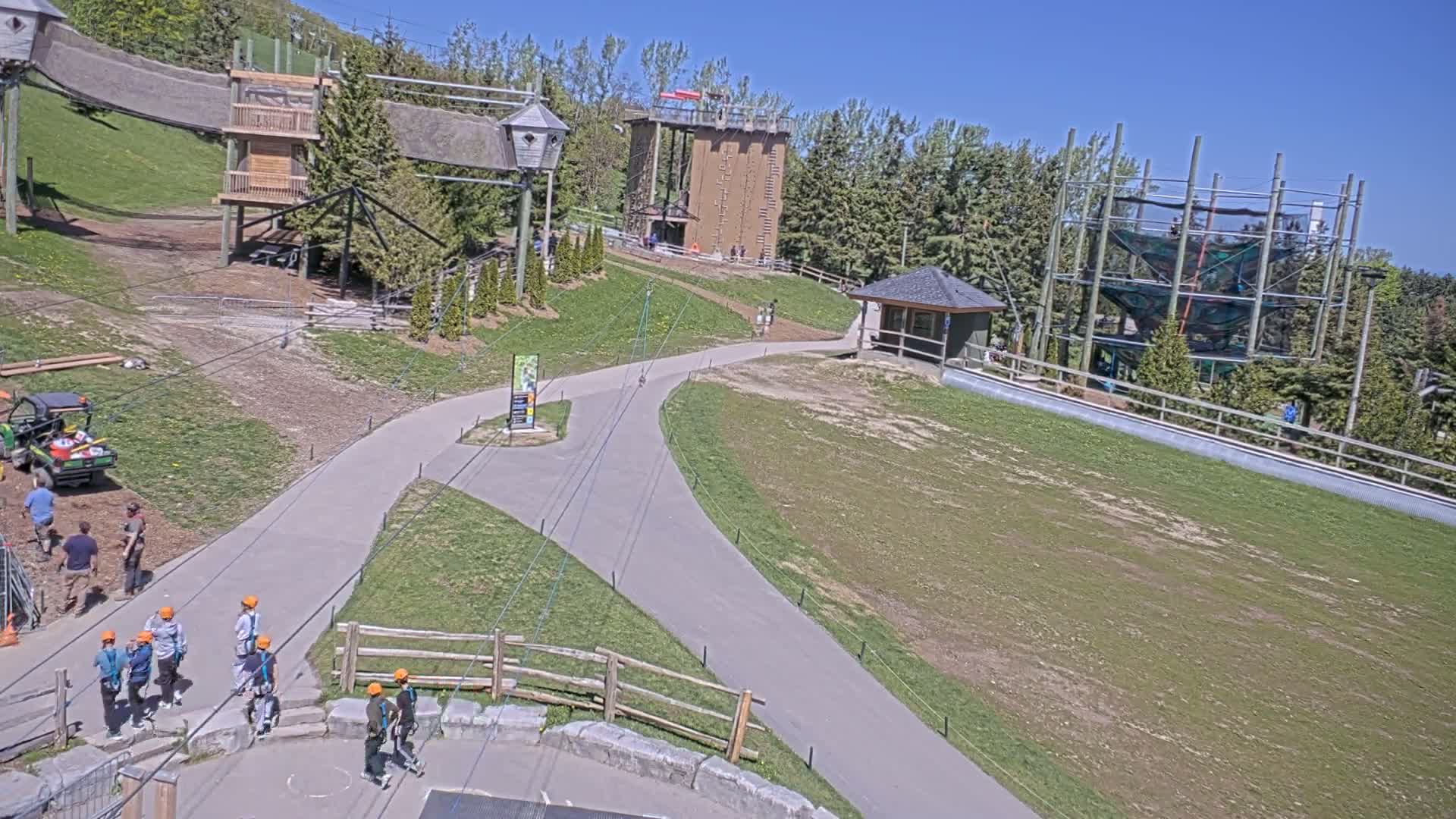 An outdoor adventure park on a sunny day features various structures, including climbing walls, zip lines, and elevated walkways, with several people present.