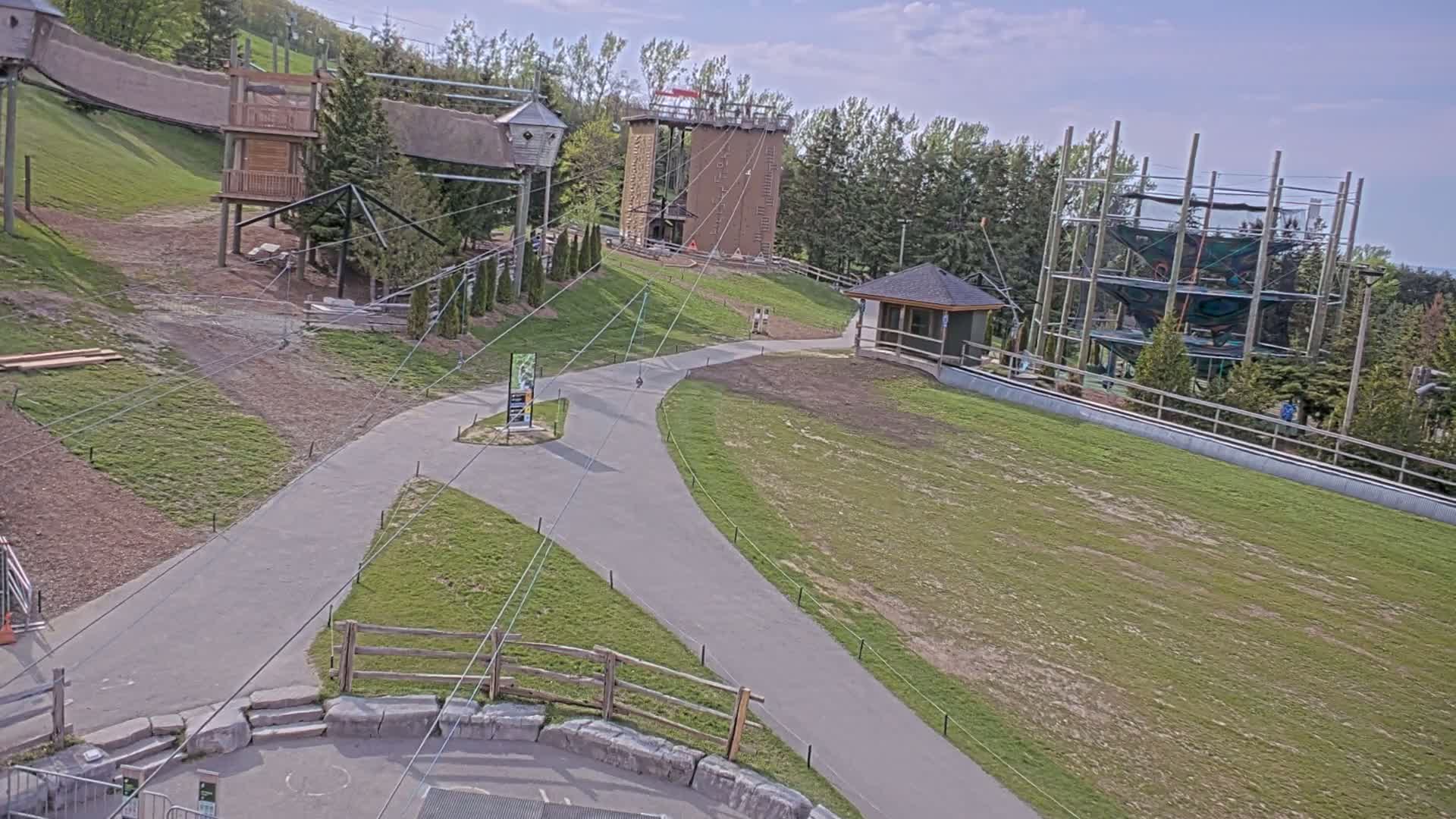 An outdoor aerial view shows a paved pathway winding through a grassy area with various wooden structures, including elevated platforms and a climbing tower, under partly cloudy skies.