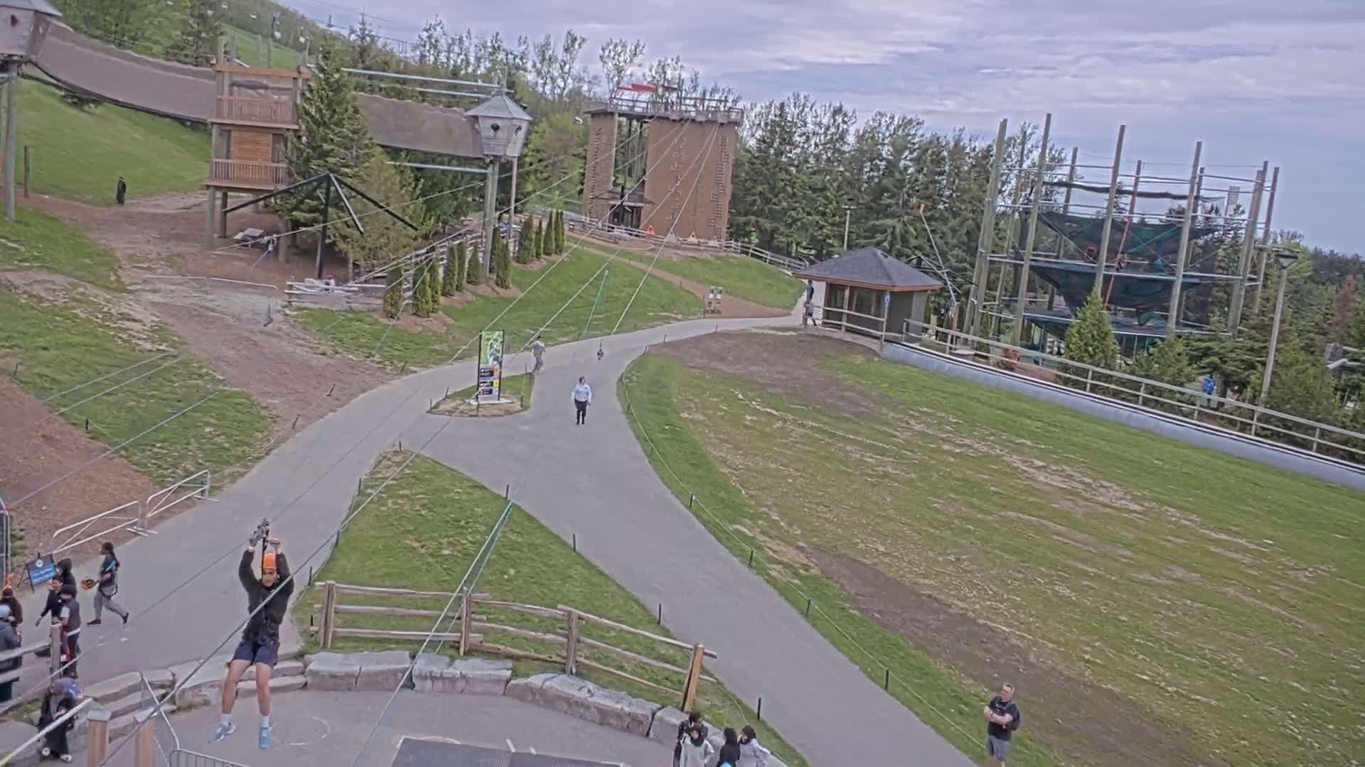 A person ziplines over a paved path in an outdoor adventure park on an overcast day, with other people walking on the paths and several structures, including climbing towers and treetop obstacle courses, visible in the background.