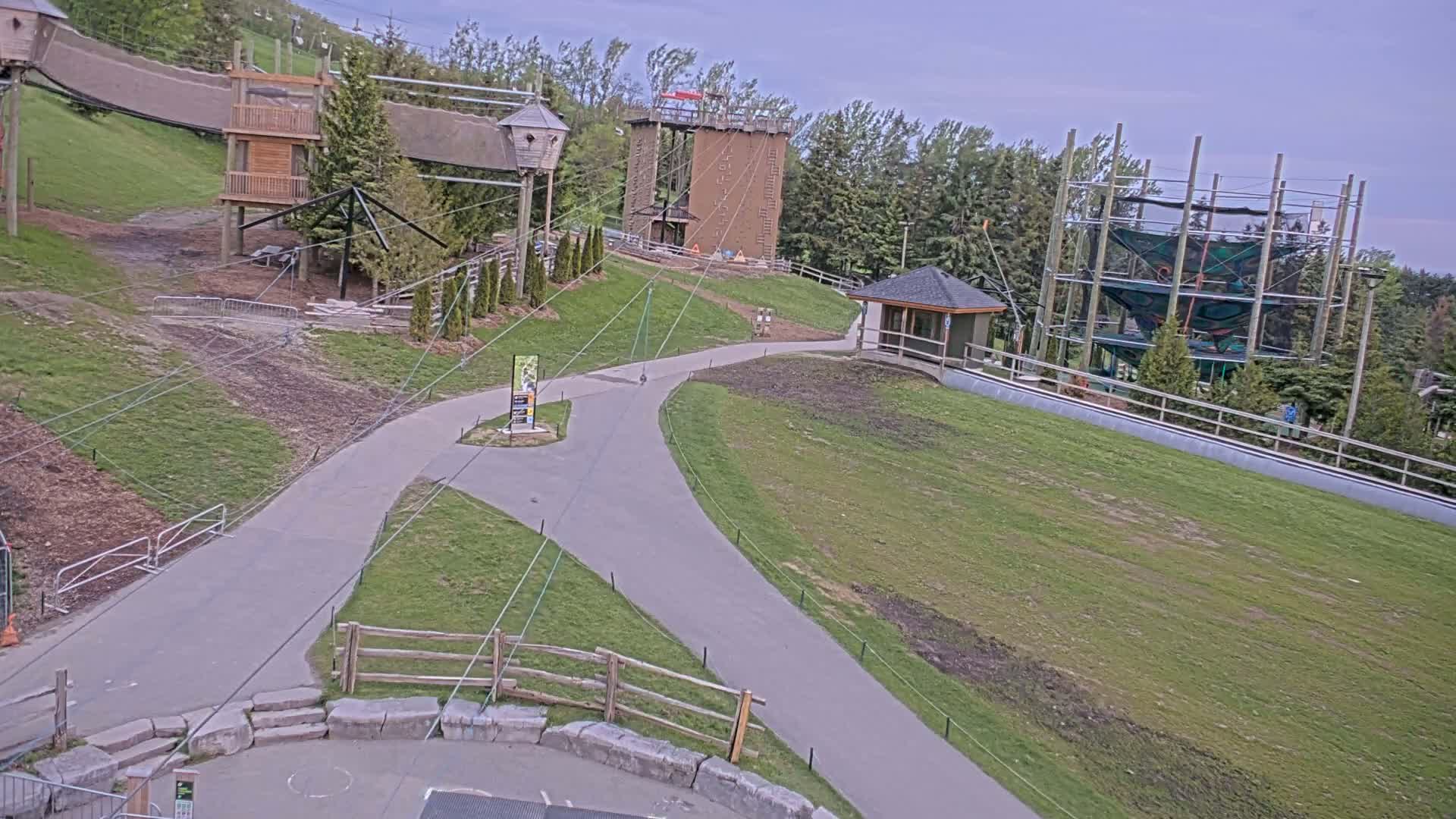 A paved pathway winds through a grassy area with various outdoor recreational structures, including climbing walls and rope courses, under an overcast sky.