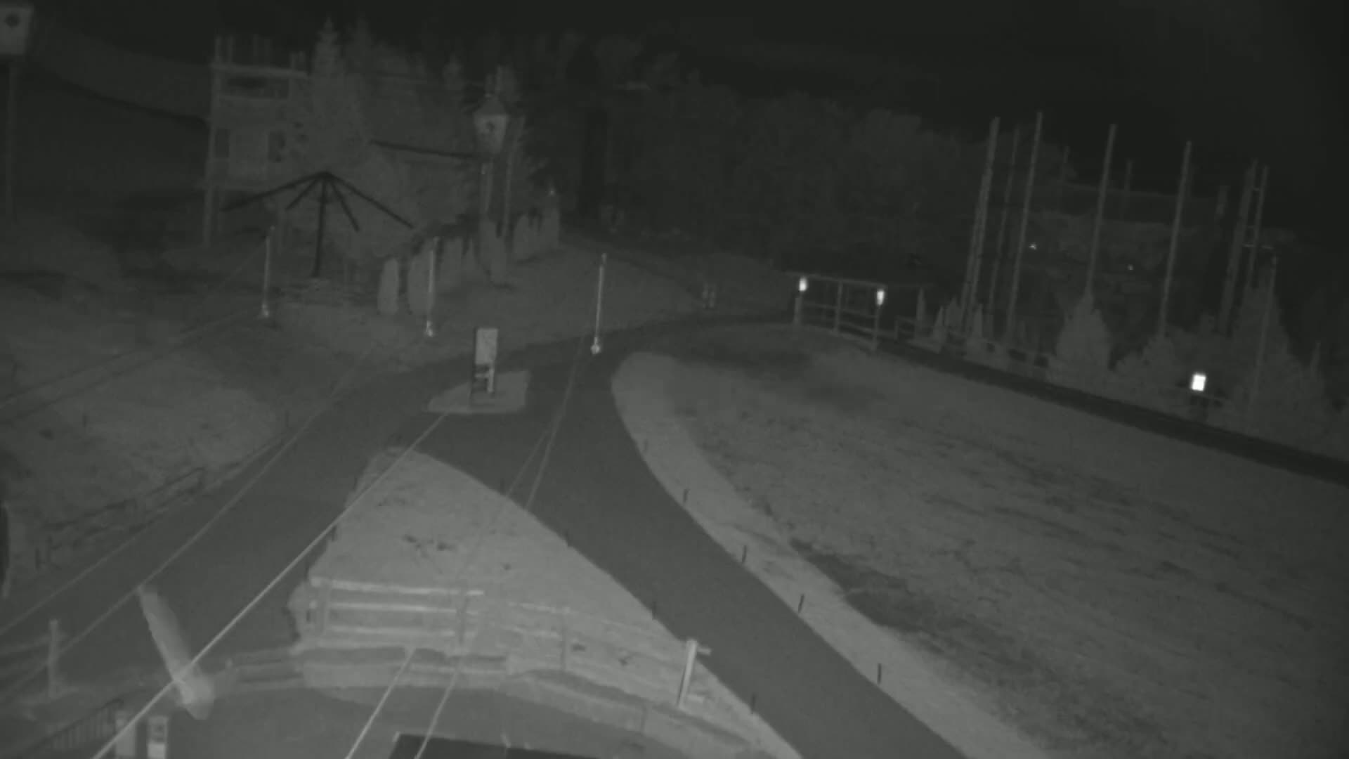 A nighttime, infrared view shows a paved intersection with a building and several tall, thin structures in the background.