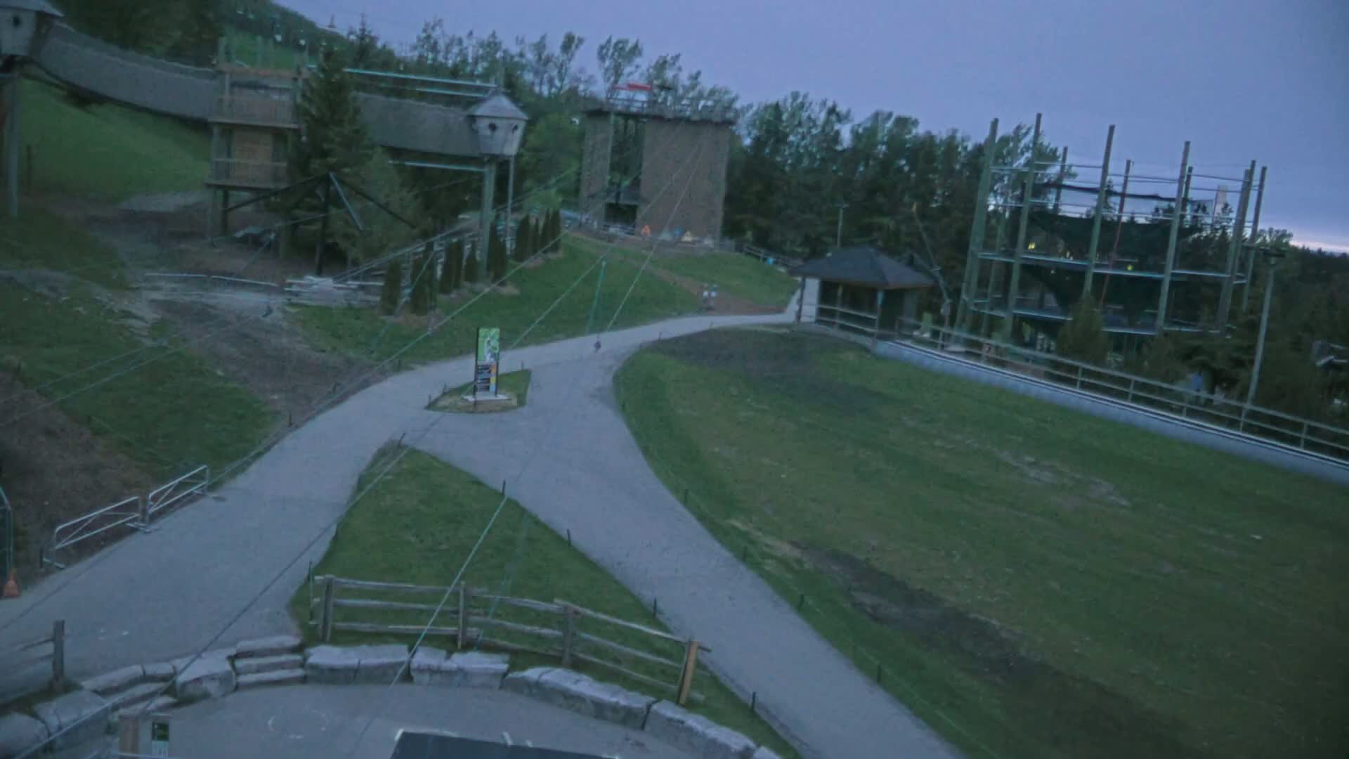 An overhead view shows a paved path winding through a grassy area with several elevated structures, including a climbing tower and treetop obstacle courses, under a dusky sky.