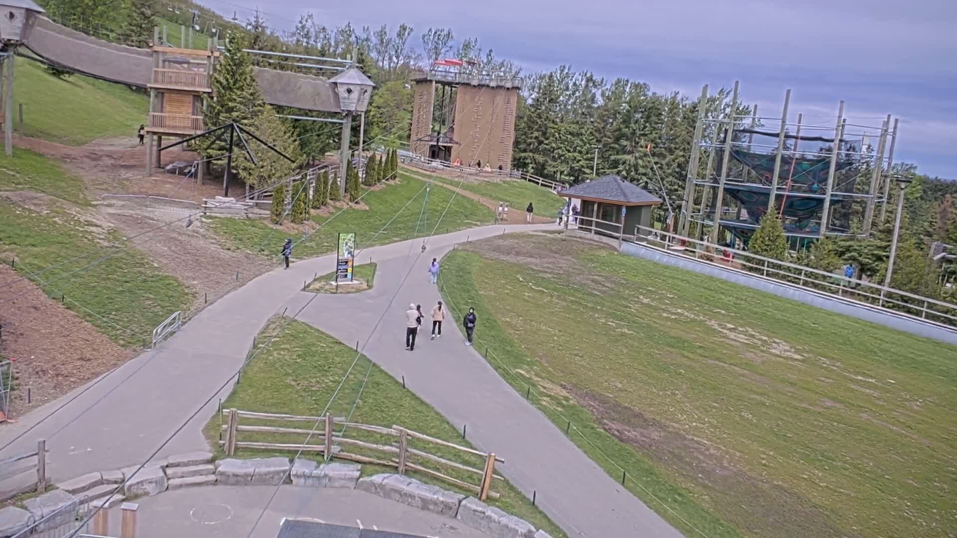 A paved pathway winds through a grassy area with several people walking, leading to various outdoor recreational structures including a climbing tower and a rope course, under a partly cloudy sky.