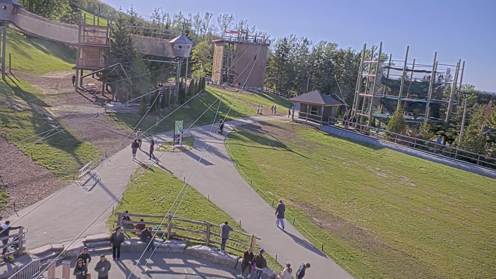 An outdoor scene on a sunny day shows a paved walkway winding through a grassy area with several wooden structures, including climbing towers and what appears to be a zip line course, with people walking and sitting in the area.
