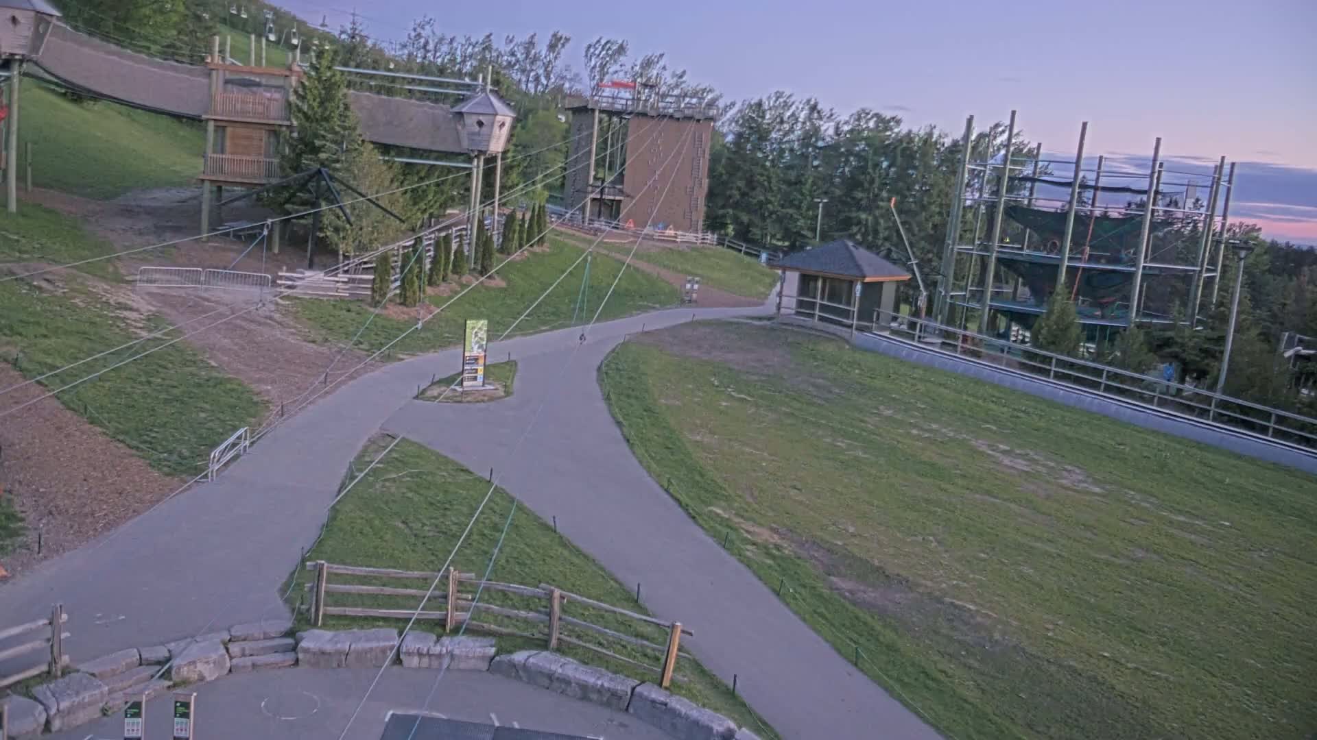 A paved pathway winds through a grassy area, past various structures including elevated treetop adventures and climbing structures, under a twilight sky.