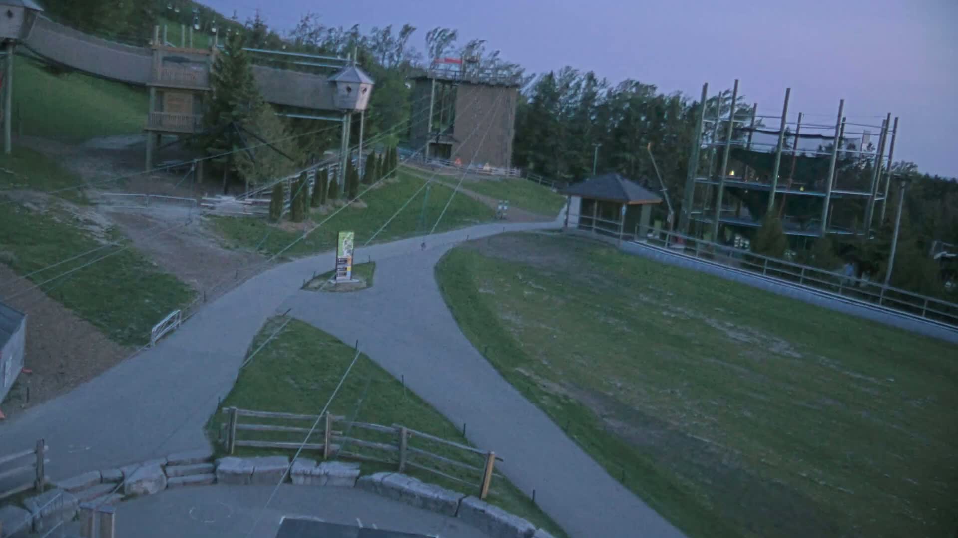 An outdoor aerial view shows a paved pathway winding through grassy areas near several elevated structures, including a climbing tower and zip-line apparatuses, under a dusky sky.