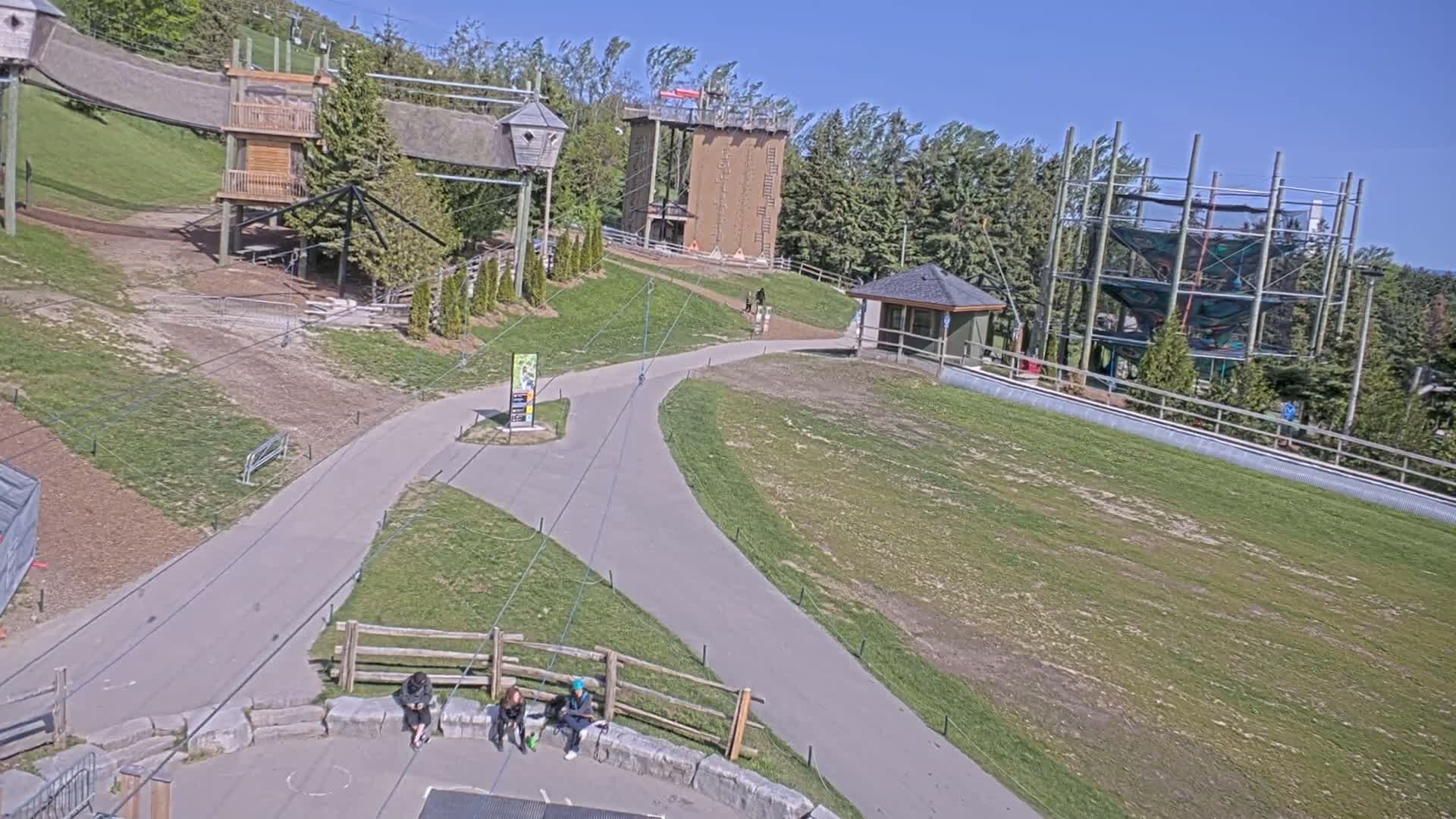 An outdoor recreational area with climbing structures, zip lines, and paved pathways on a sunny day with a light breeze.