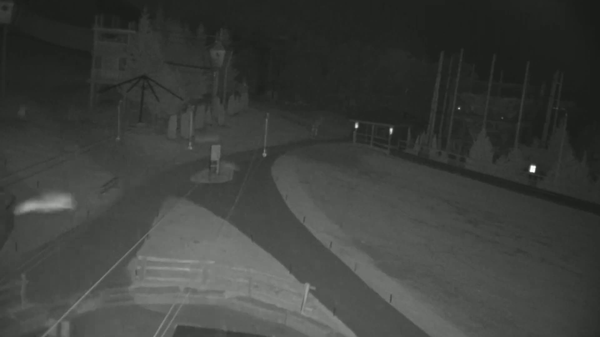 A nighttime, low-light view shows a paved road curving through a snow-covered area with some structures and trees in the background.