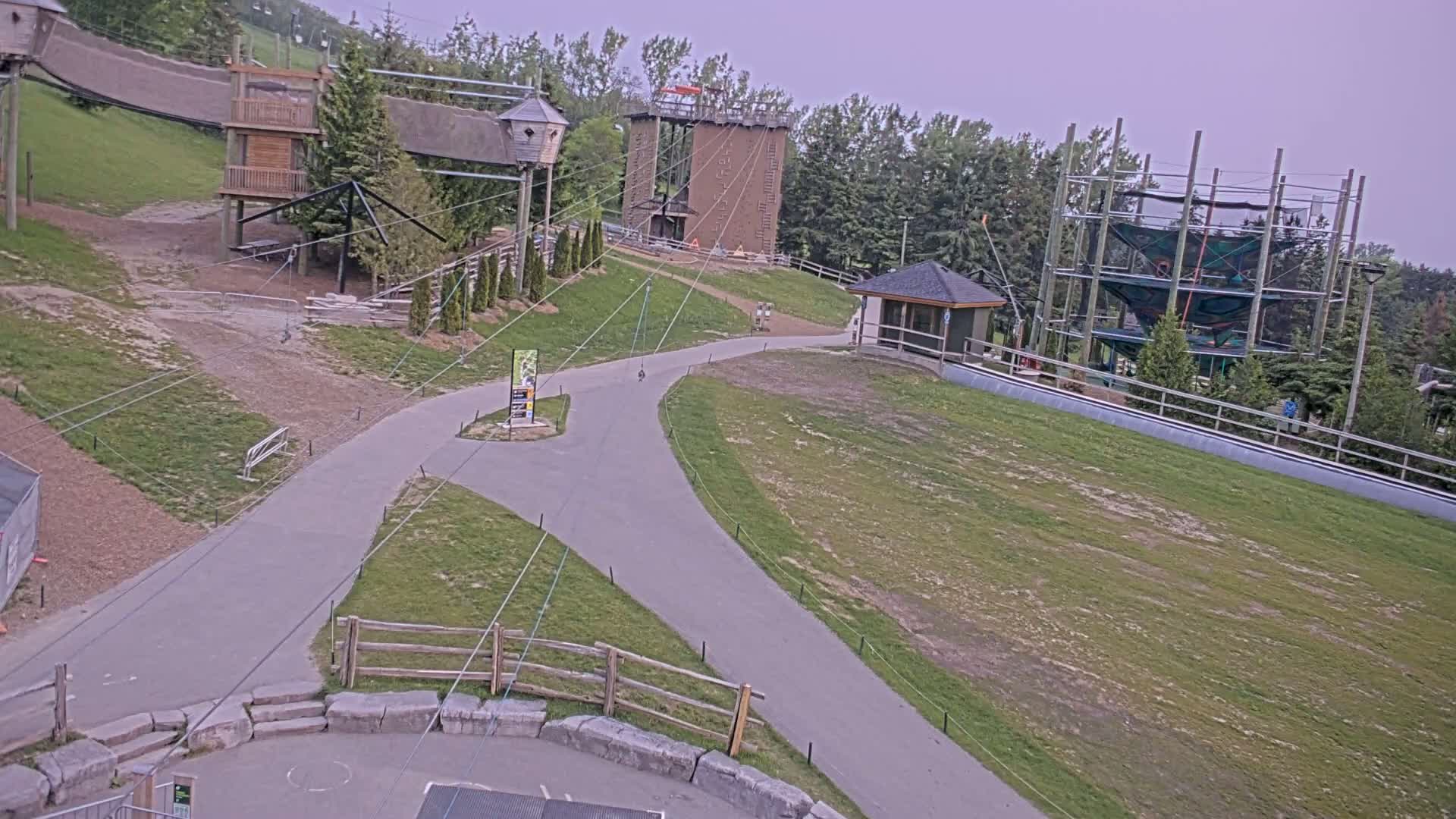 An outdoor paved pathway winds through a grassy area with various structures including climbing towers, tree houses, and a small building, under an overcast sky.