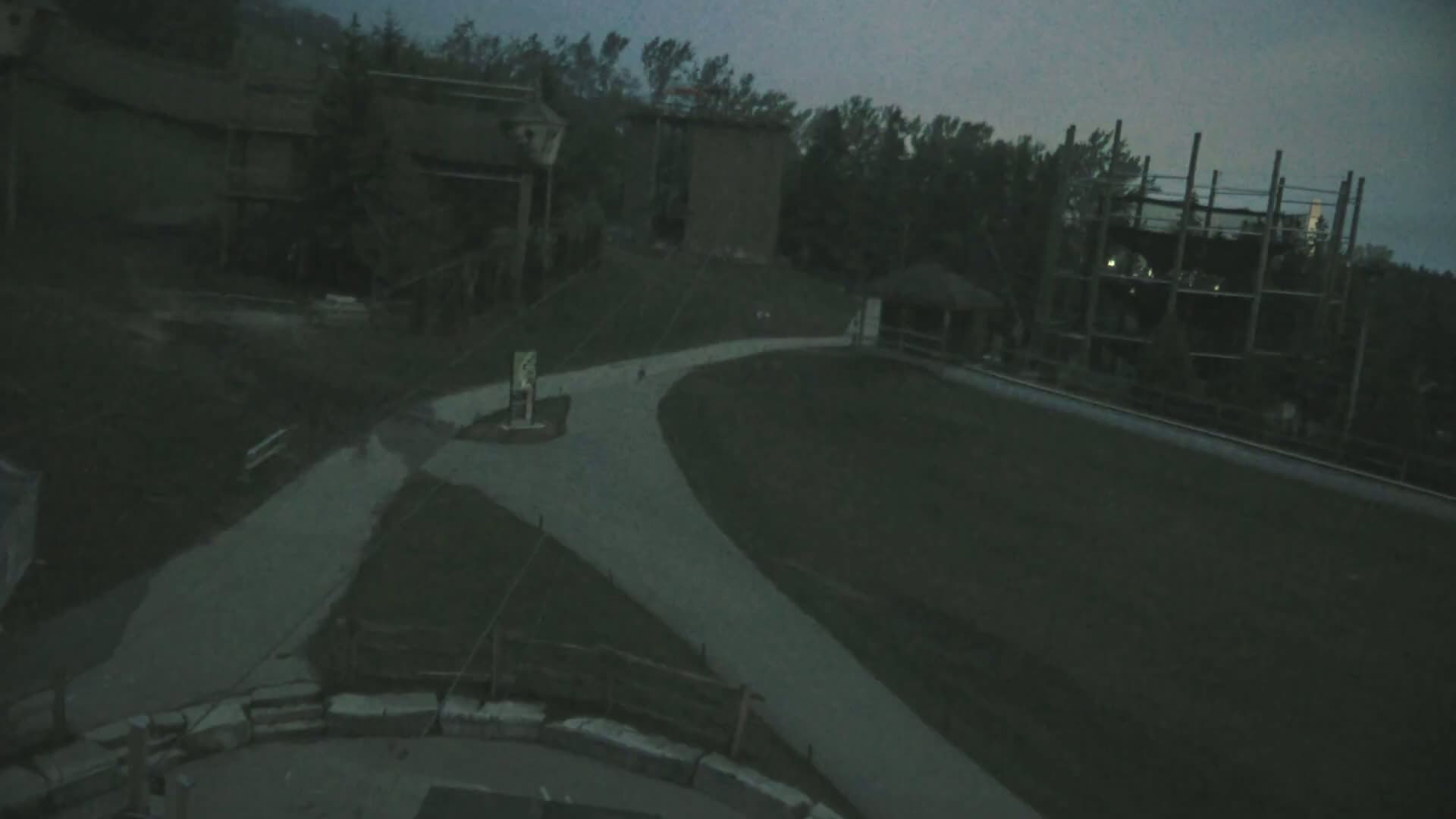 A paved pathway curves through a dimly lit outdoor area with buildings and trees under a dark, overcast sky.
