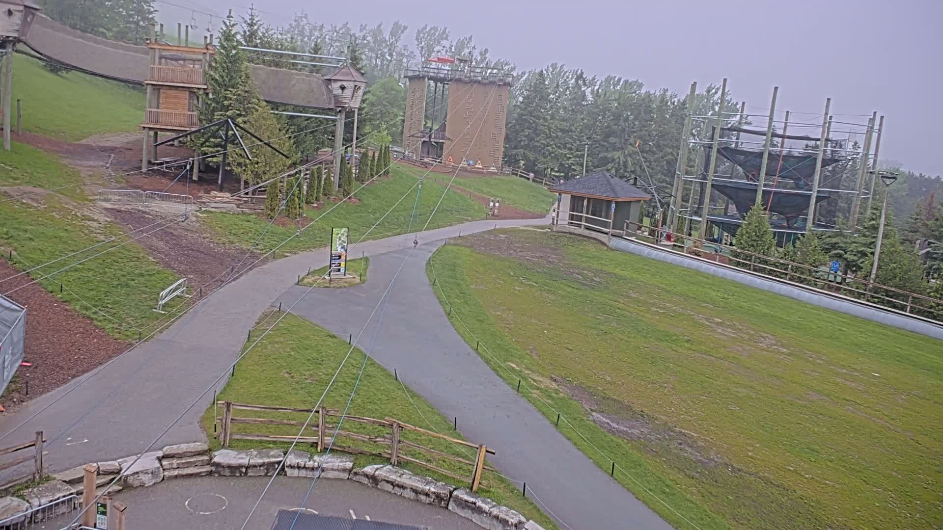 A paved pathway winds through a grassy area, past various outdoor recreational structures including a climbing wall, rope courses, and a small building, all under overcast skies.