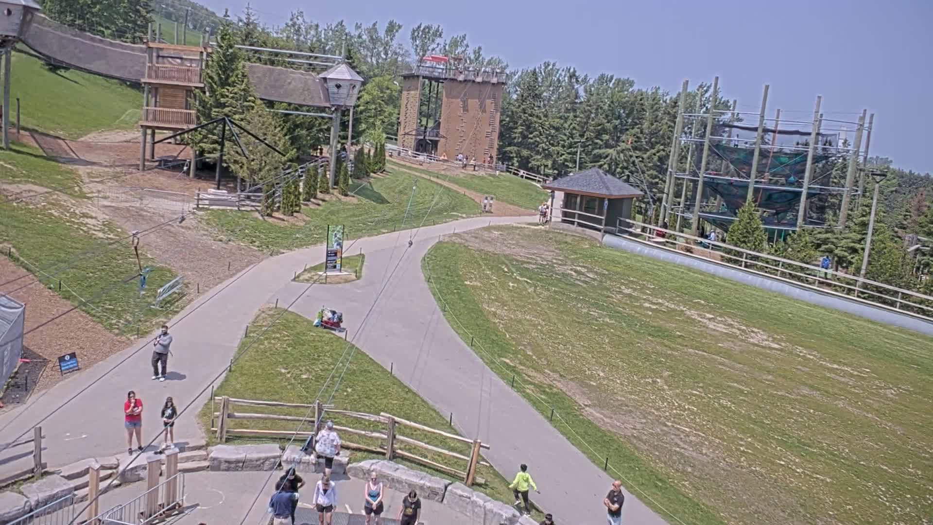 An outdoor recreation area with climbing structures, zip lines, and paved paths, under sunny skies.