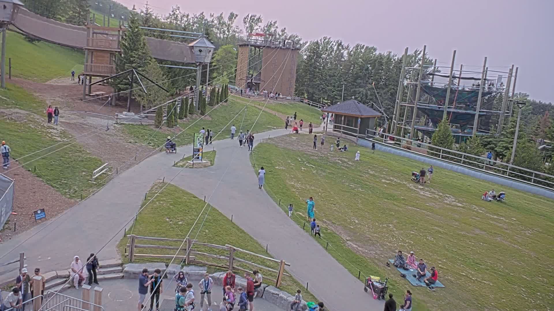 An outdoor recreational area with several people walking on paved paths and relaxing on grassy areas, near various playground structures and climbing towers, under an overcast sky.