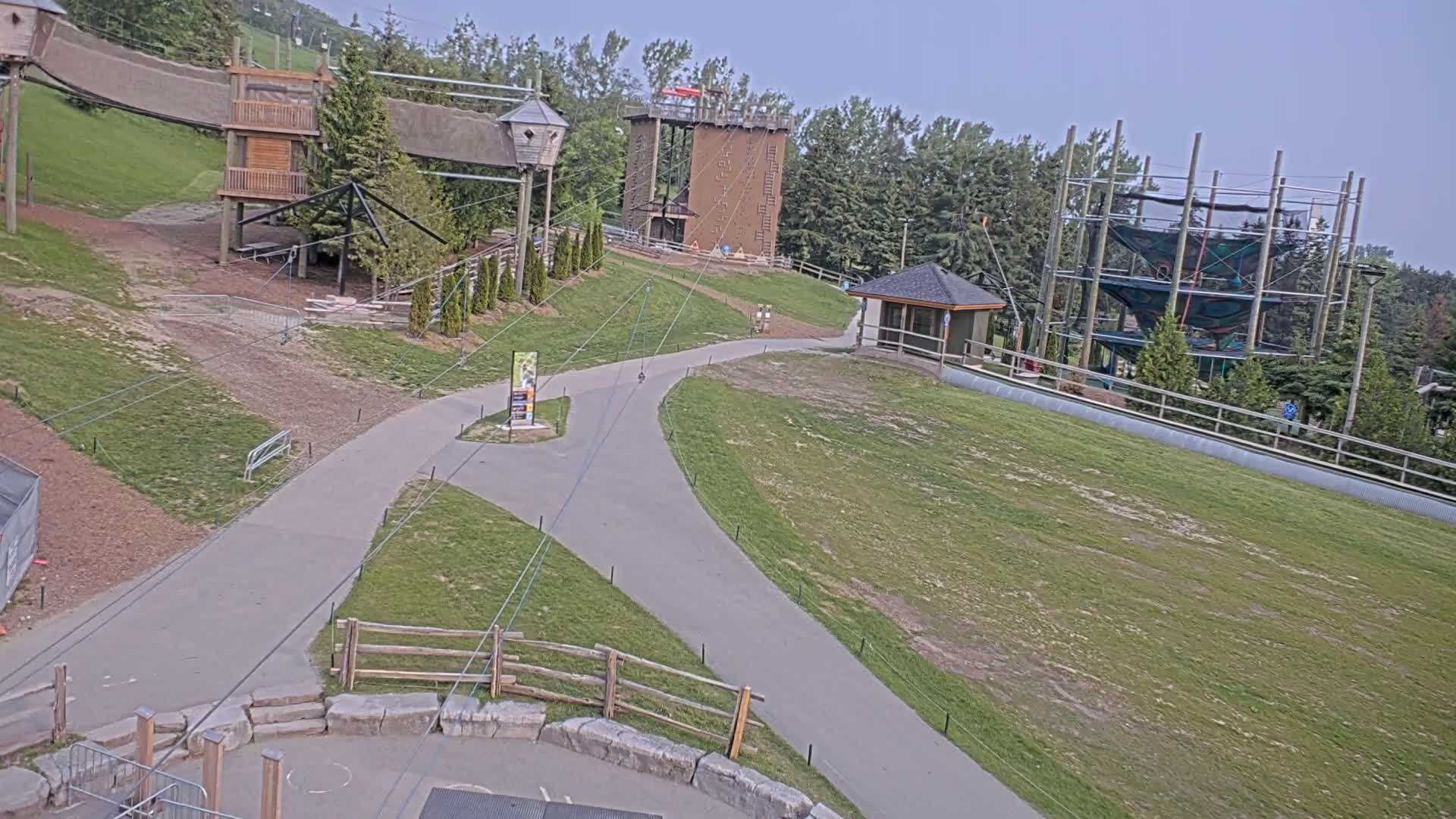 A paved pathway winds through a grassy area with various elevated play structures, including climbing walls and nets, under a mostly clear sky.