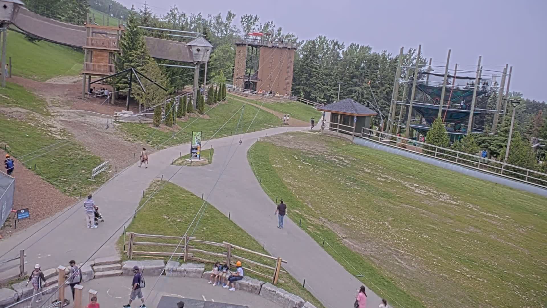 An outdoor recreational area with various play structures, including a climbing wall and ropes course, is seen on an overcast day.