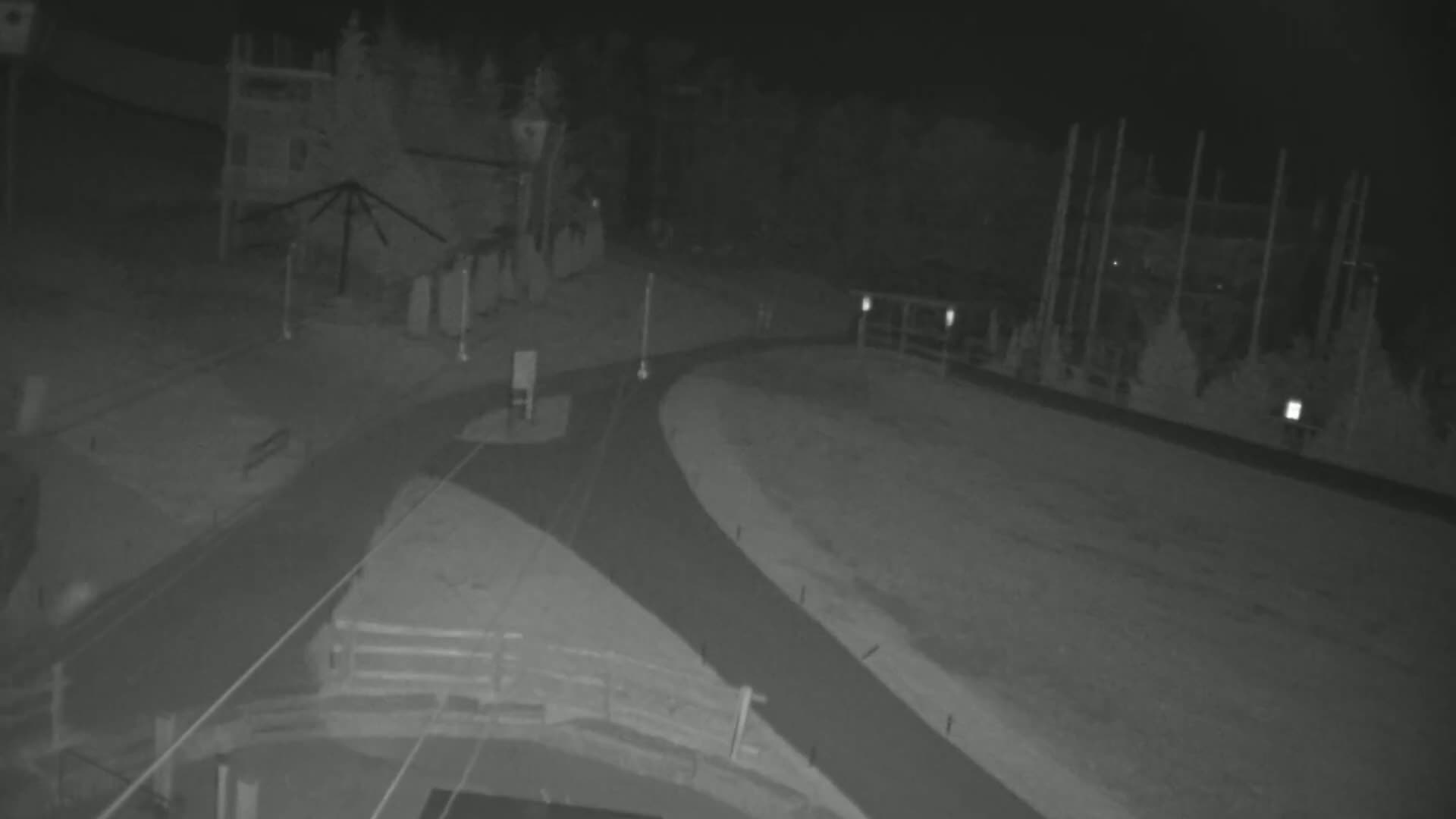A nighttime, high-angle view shows a paved roadway curving through a snow-covered area near buildings and trees.