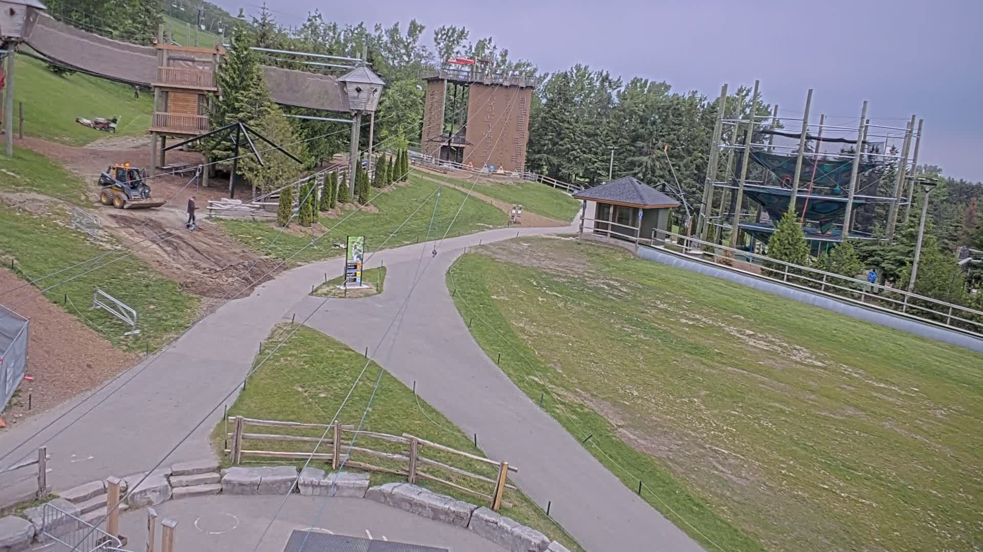 An outdoor scene on an overcast day shows a paved path winding through a grassy area with various structures, including climbing walls, rope courses, and a small building, with some construction or landscaping work underway.