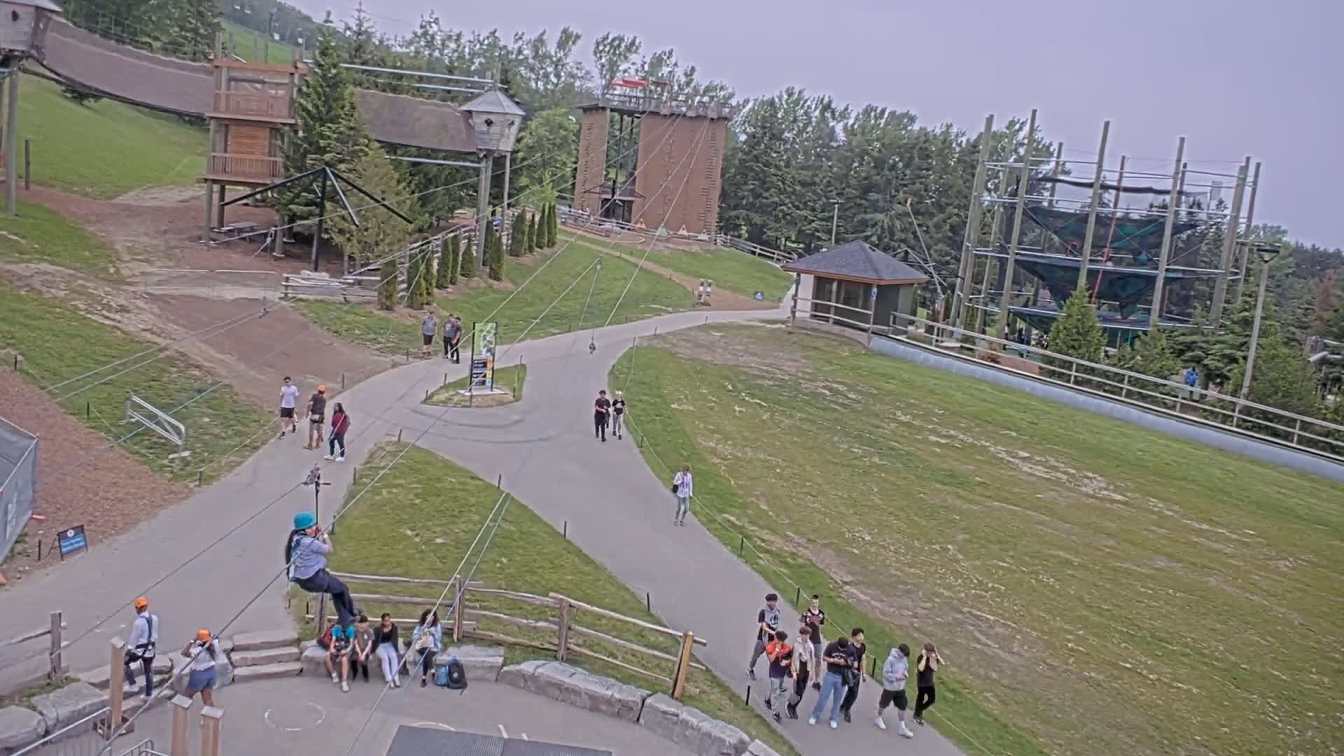 An outdoor recreational area with zip lines, climbing structures, and walking paths features several people walking and engaging in activities under an overcast sky.