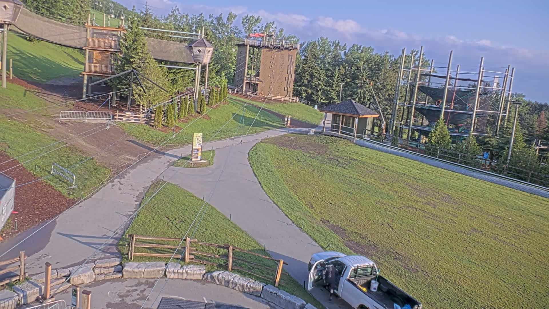 An aerial view shows a paved pathway winding through a grassy area with several wooden structures, including a climbing tower and a ropes course, under a mostly clear sky.