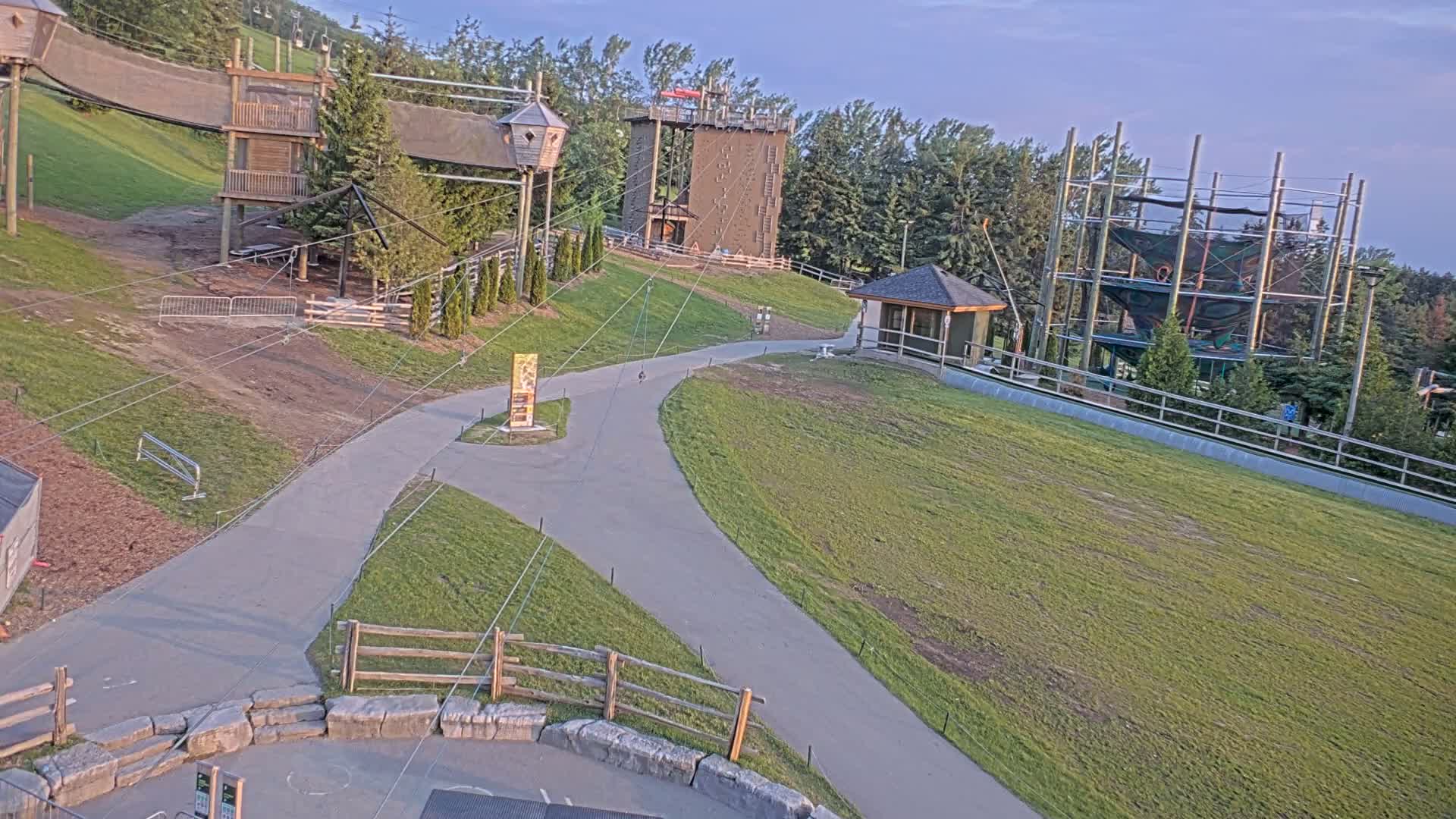 A paved pathway winds through a grassy area with various outdoor adventure structures, including climbing towers and a zip line course, under a clear evening sky.