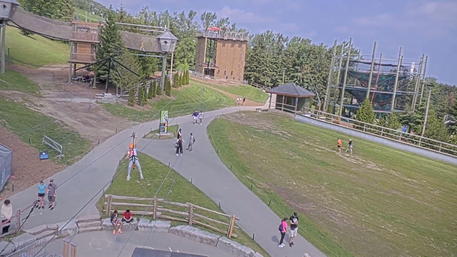 An outdoor adventure park on a sunny day features several people walking on paved paths, others zip-lining, and various climbing structures and obstacles.