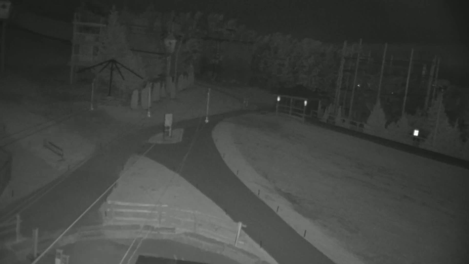 A nighttime, low-light view shows a paved path curving through a snow-covered area near some trees and structures.