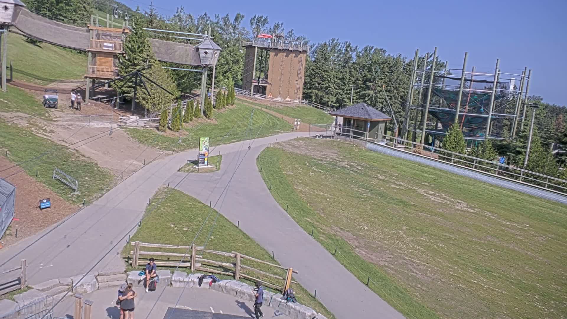 A paved pathway winds through a grassy area with various outdoor adventure structures, including climbing walls and rope courses, under a clear sunny sky.