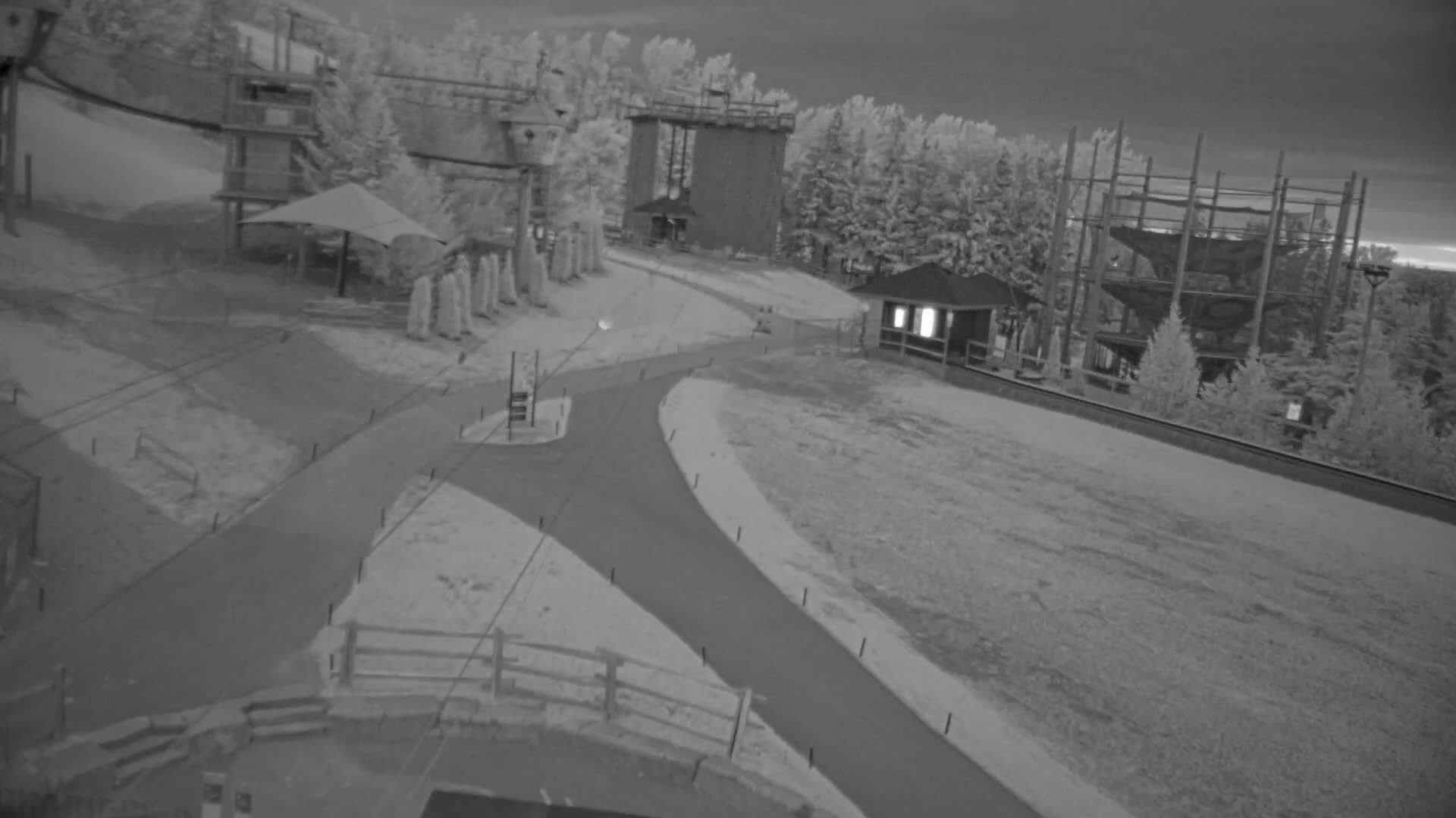 An aerial view of a snowy outdoor area shows paved pathways winding between a playground structure, a tall tower, a small building, and a large climbing structure under a dark, overcast sky.