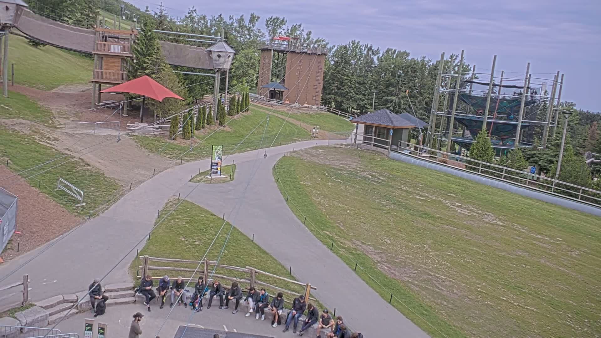 An outdoor scene on an overcast day shows a paved path winding through a grassy area with several wooden structures, including climbing walls and a multi-level ropes course, and a group of people sitting on a stone bench.