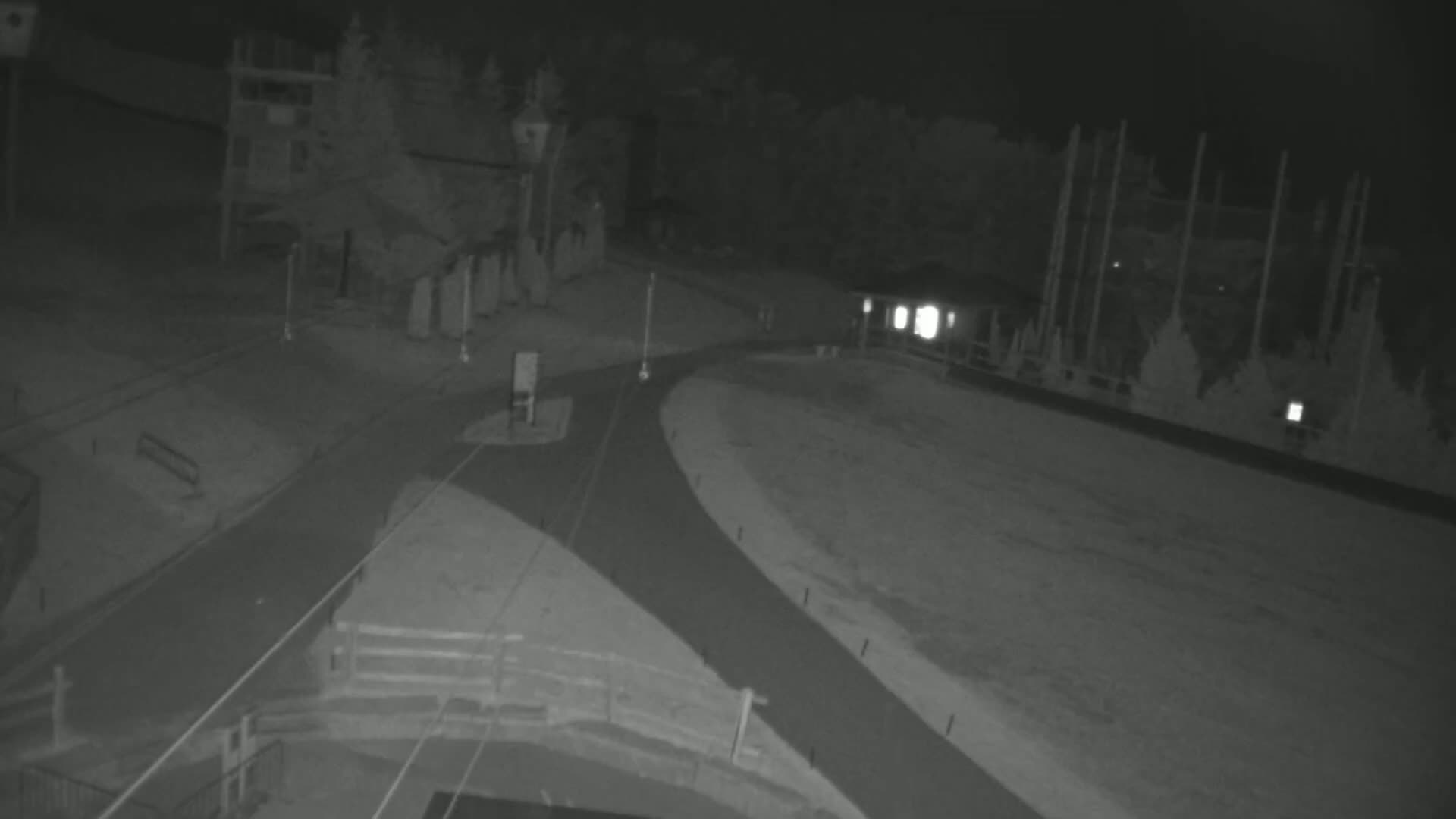 A nighttime, overhead view of a paved area with intersecting paths, some buildings, and trees, under dark conditions.