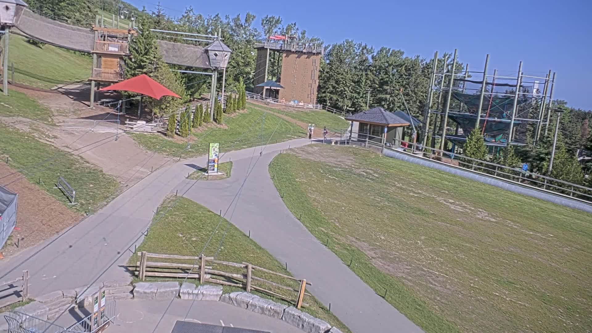 A paved pathway winds through a grassy area with various outdoor recreational structures, including climbing towers and a ropes course, under a clear sunny sky.