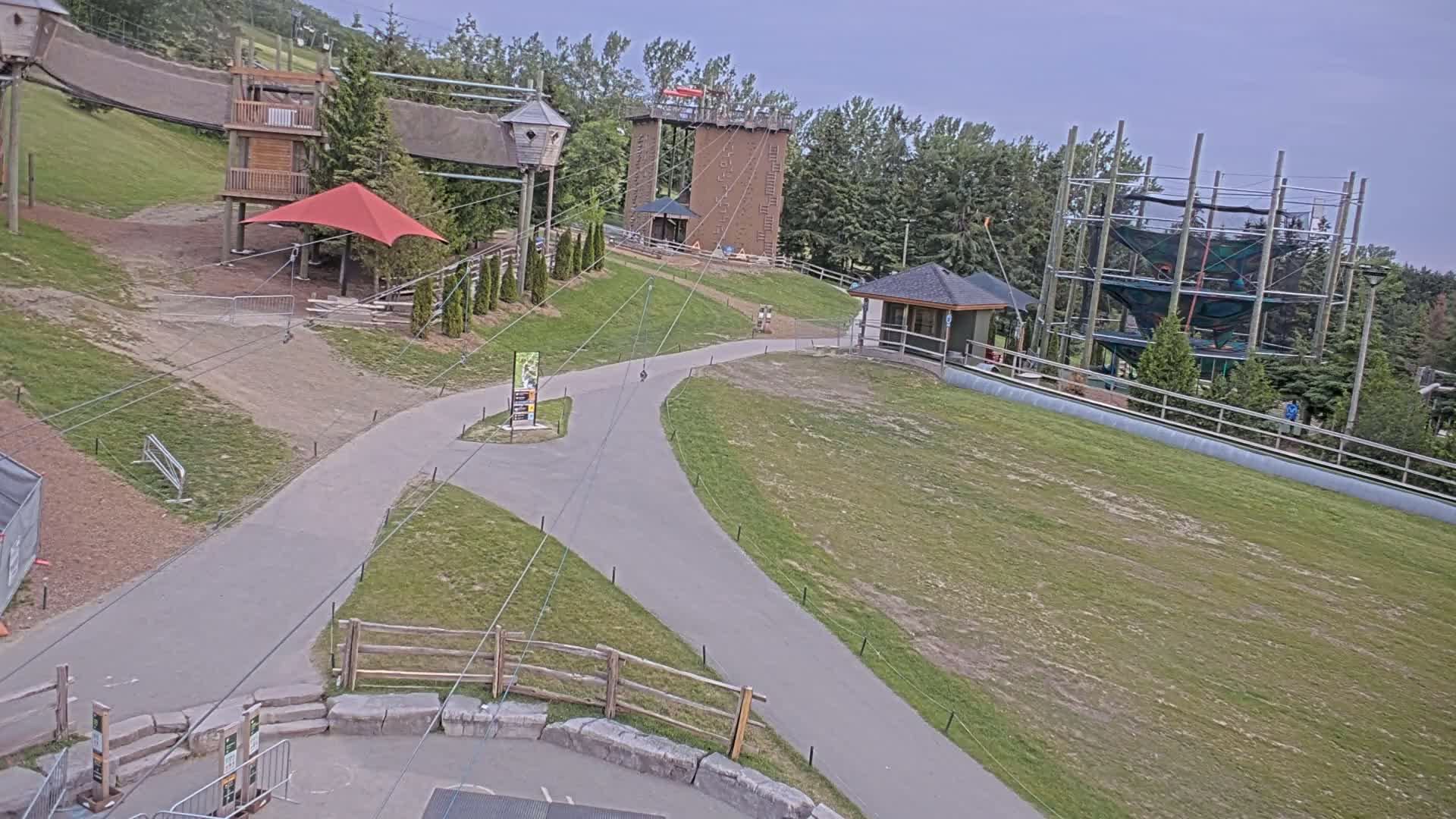 An outdoor area with paved paths, several wooden structures including a climbing wall and a ropes course, and a small building under an overcast sky.