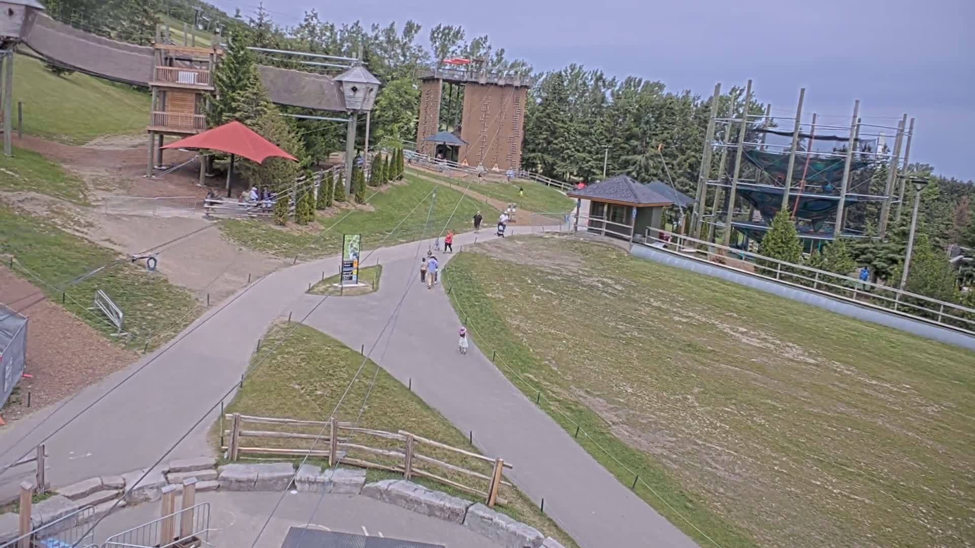 A paved path winds through a grassy area with several playground structures, including climbing walls and rope courses, under an overcast sky.