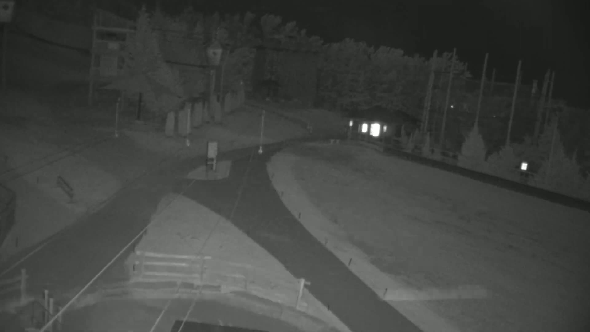 A nighttime, low-light view shows a paved road splitting into two directions, surrounded by trees and a small building.