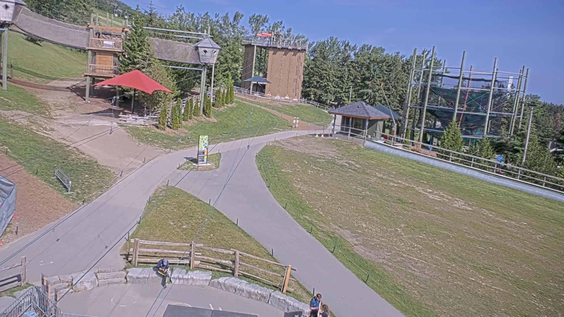 A paved pathway winds through a grassy area with various outdoor recreational structures, including climbing towers and a zip line course, under a sunny sky.