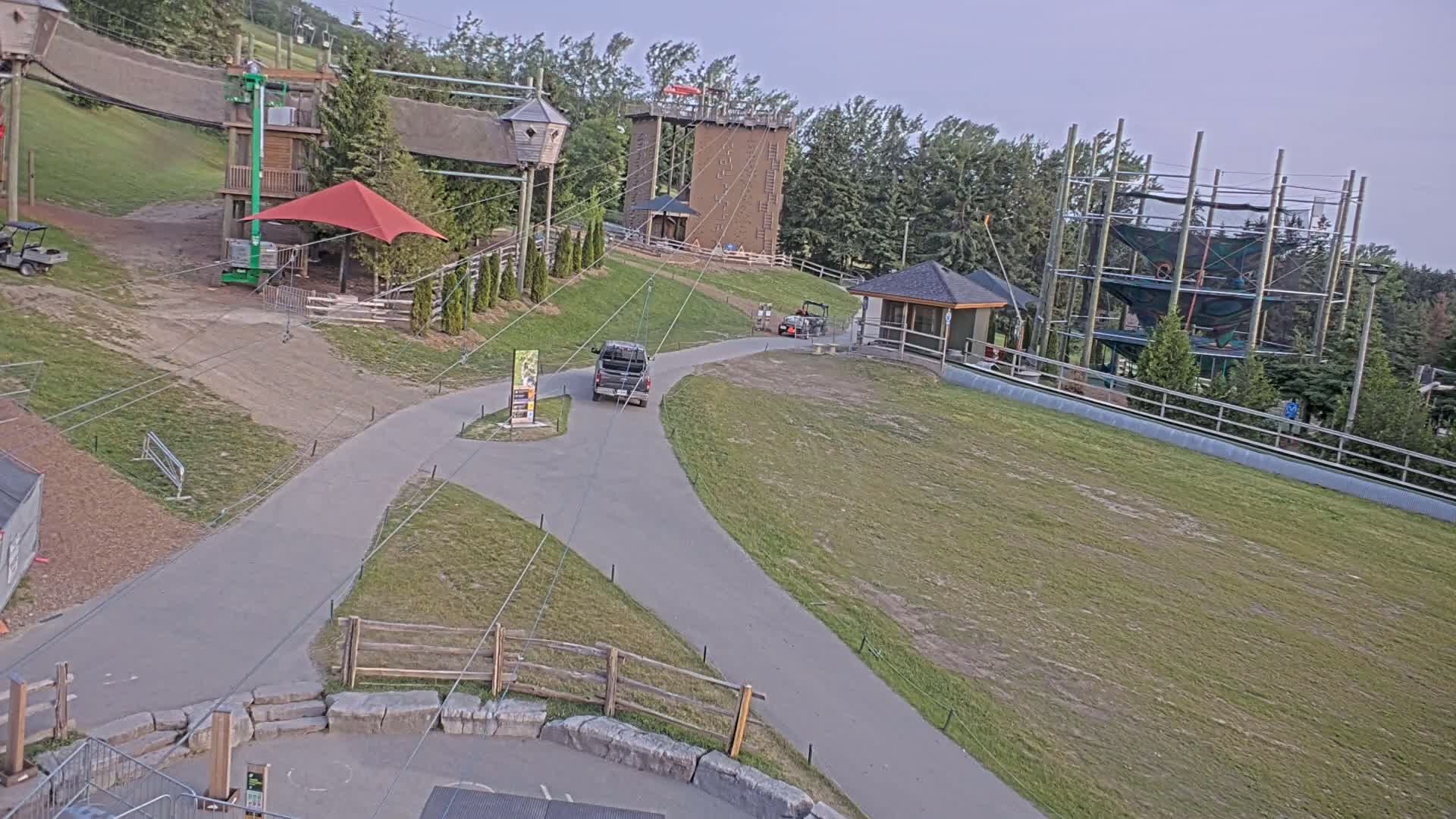 An aerial view shows a paved path winding through a grassy area with various outdoor recreational structures, including climbing walls and a ropes course, under a partly cloudy sky.