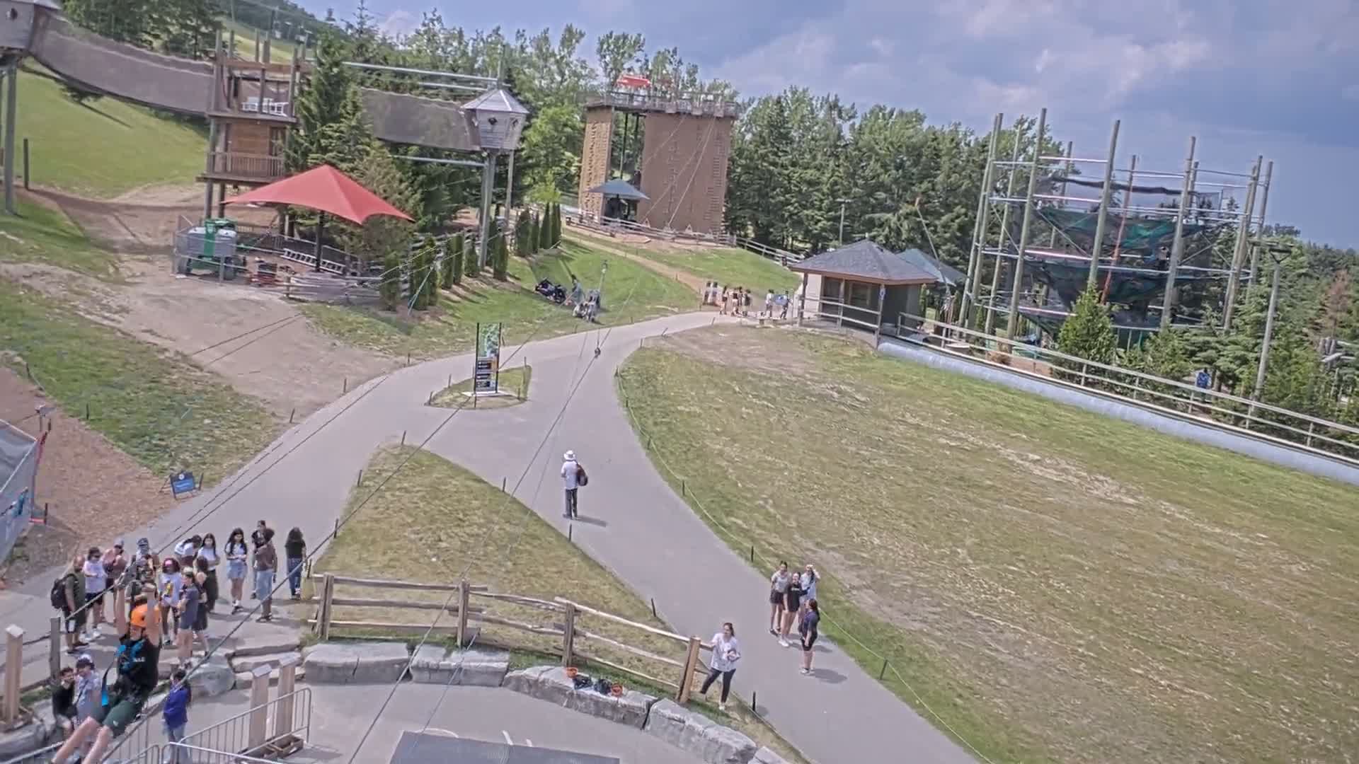 An outdoor recreational area with climbing structures, a zip line, and several people, under partly cloudy skies.