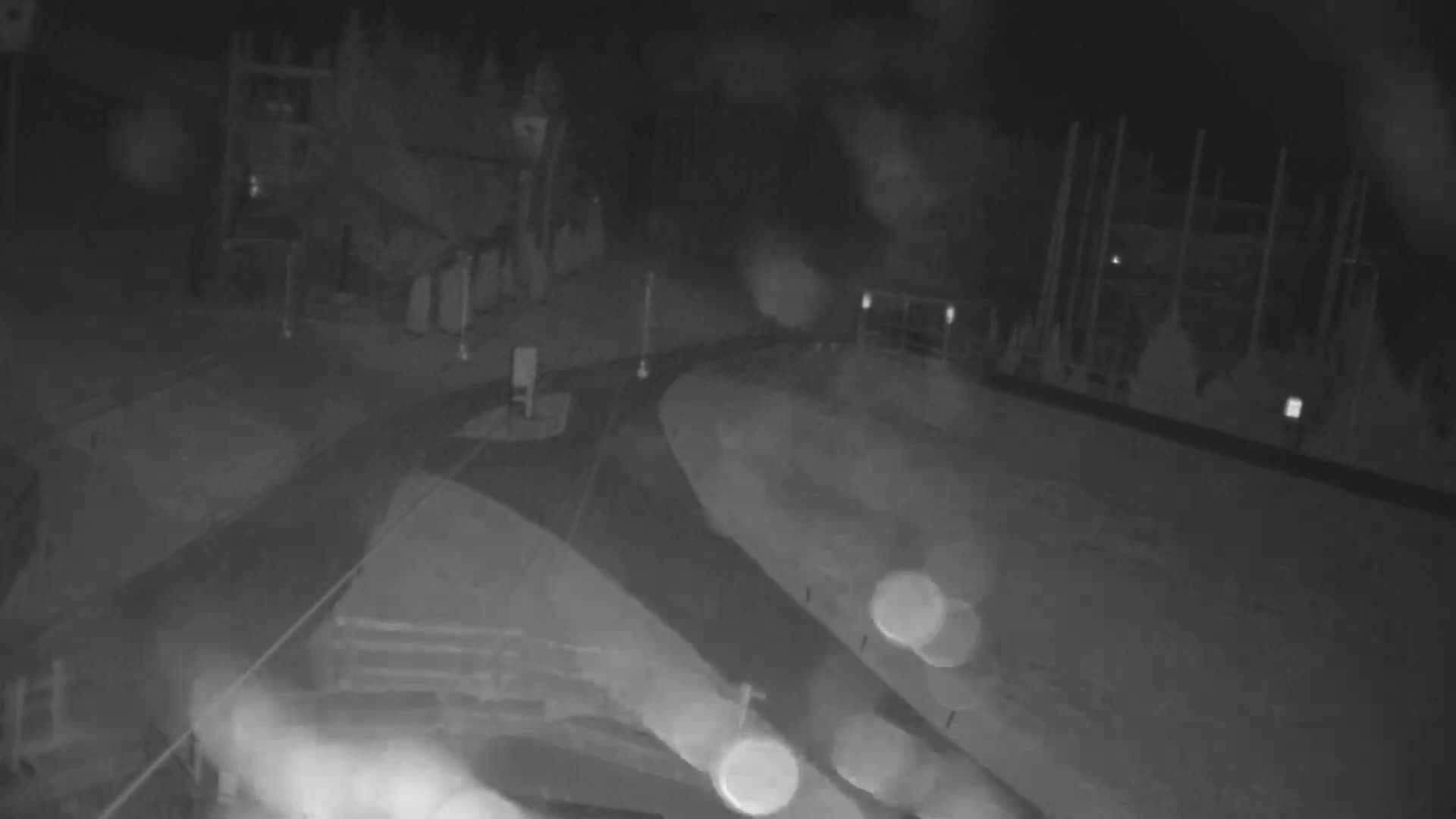 A nighttime, rain-streaked view of a dark roadway intersection with indistinct structures and blurry lights in the background.