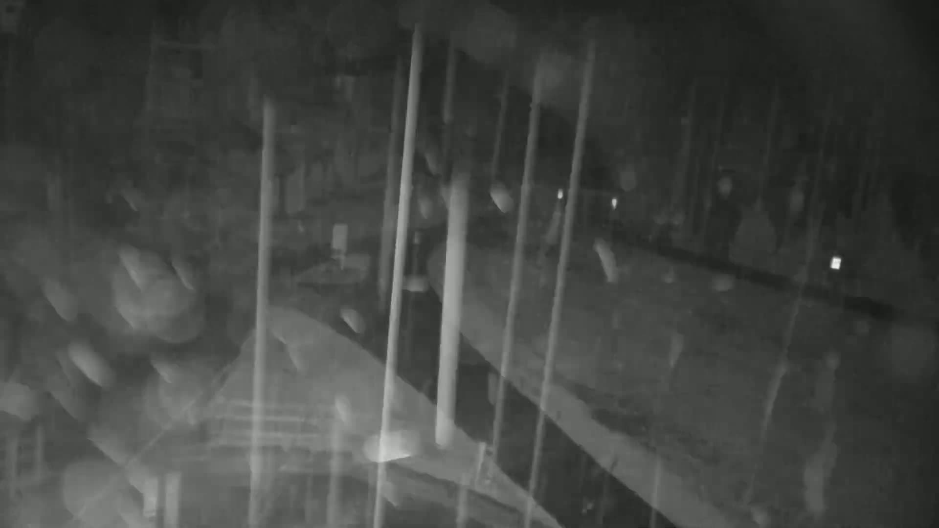 A nighttime, blurry, low-light image shows rain falling on a mostly empty outdoor area with several vertical poles or structures visible.