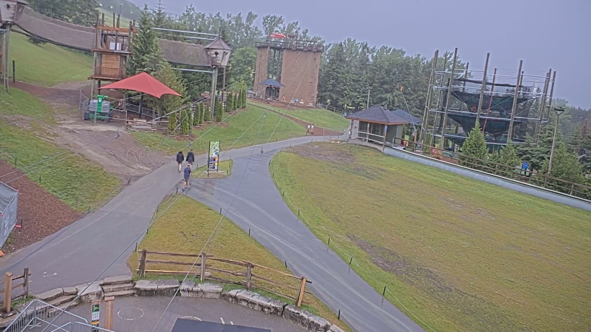 An outdoor recreational area with paved pathways, several structures including a climbing tower and ropes course, and a few people walking, all under an overcast sky.