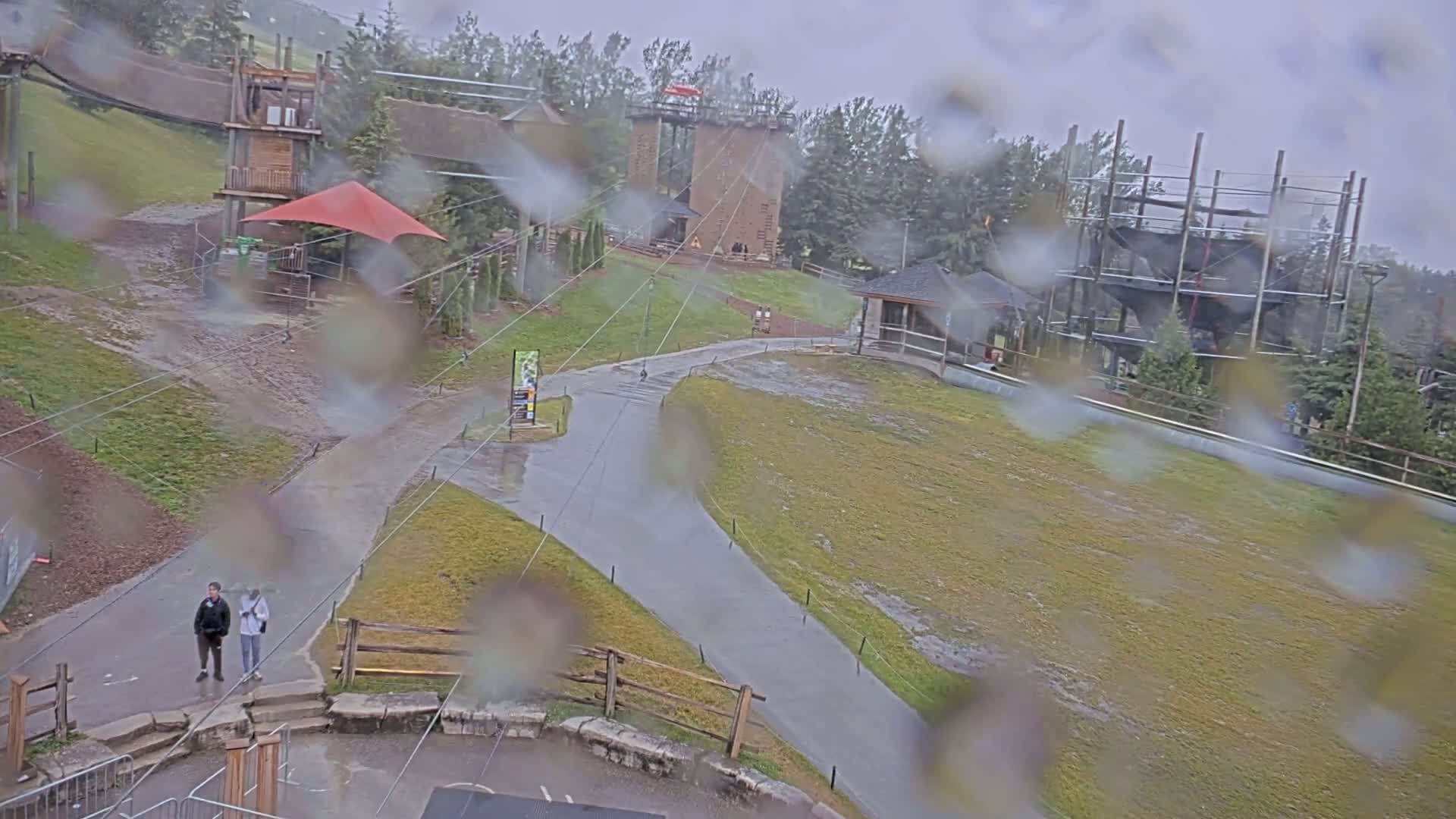 A rain-soaked outdoor recreational area with various structures, including a climbing tower, zip line course, and covered pavilion, is seen with two people standing near a walkway.