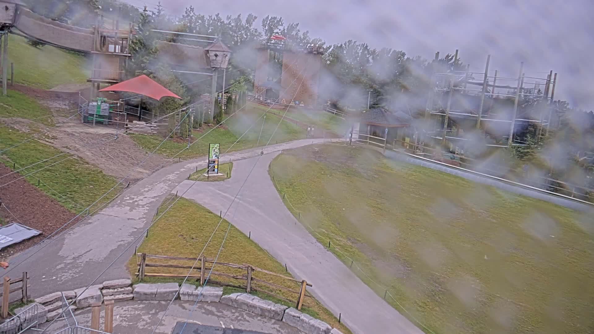 A paved path winds through a grassy area with various outdoor structures, including a climbing wall and play areas, all viewed through a rain-streaked window.