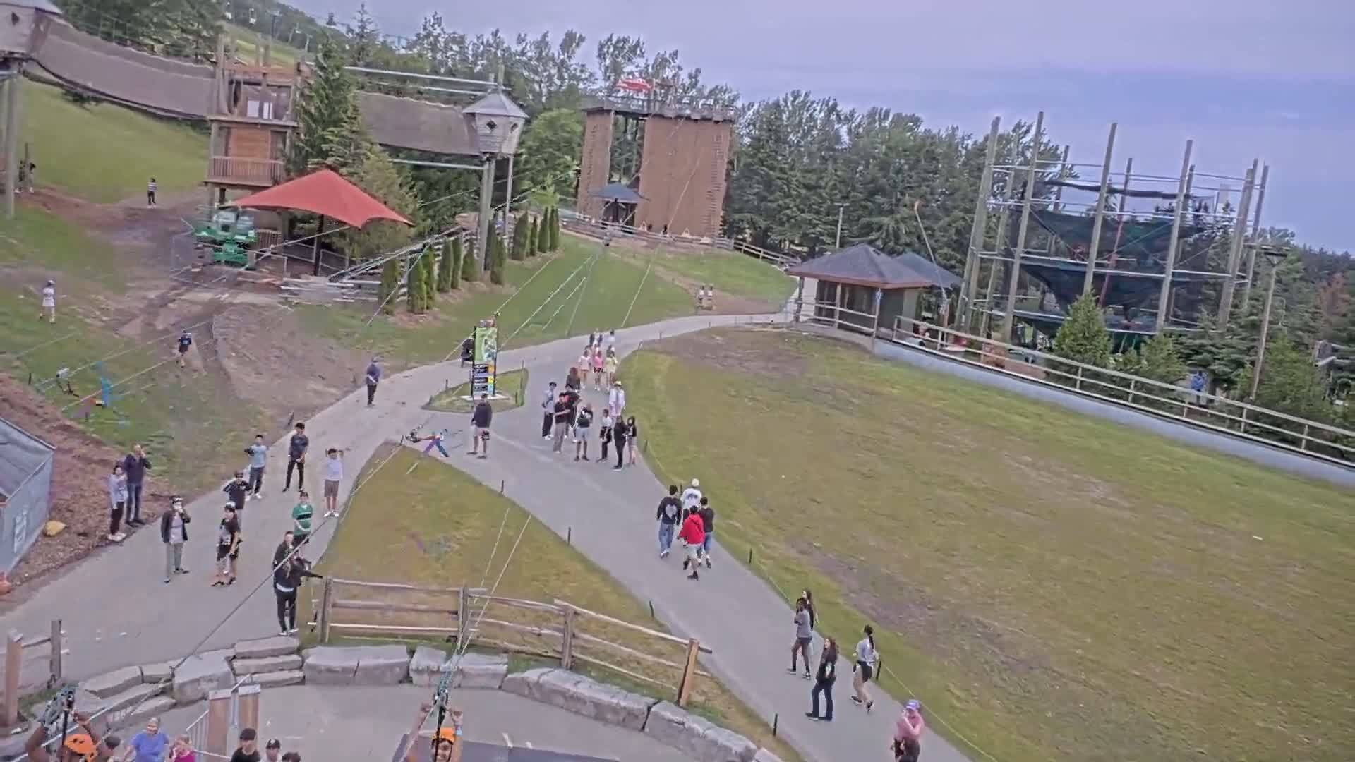An outdoor amusement park on a partly cloudy day features people walking on paved paths near wooden structures, climbing walls, and zip lines.