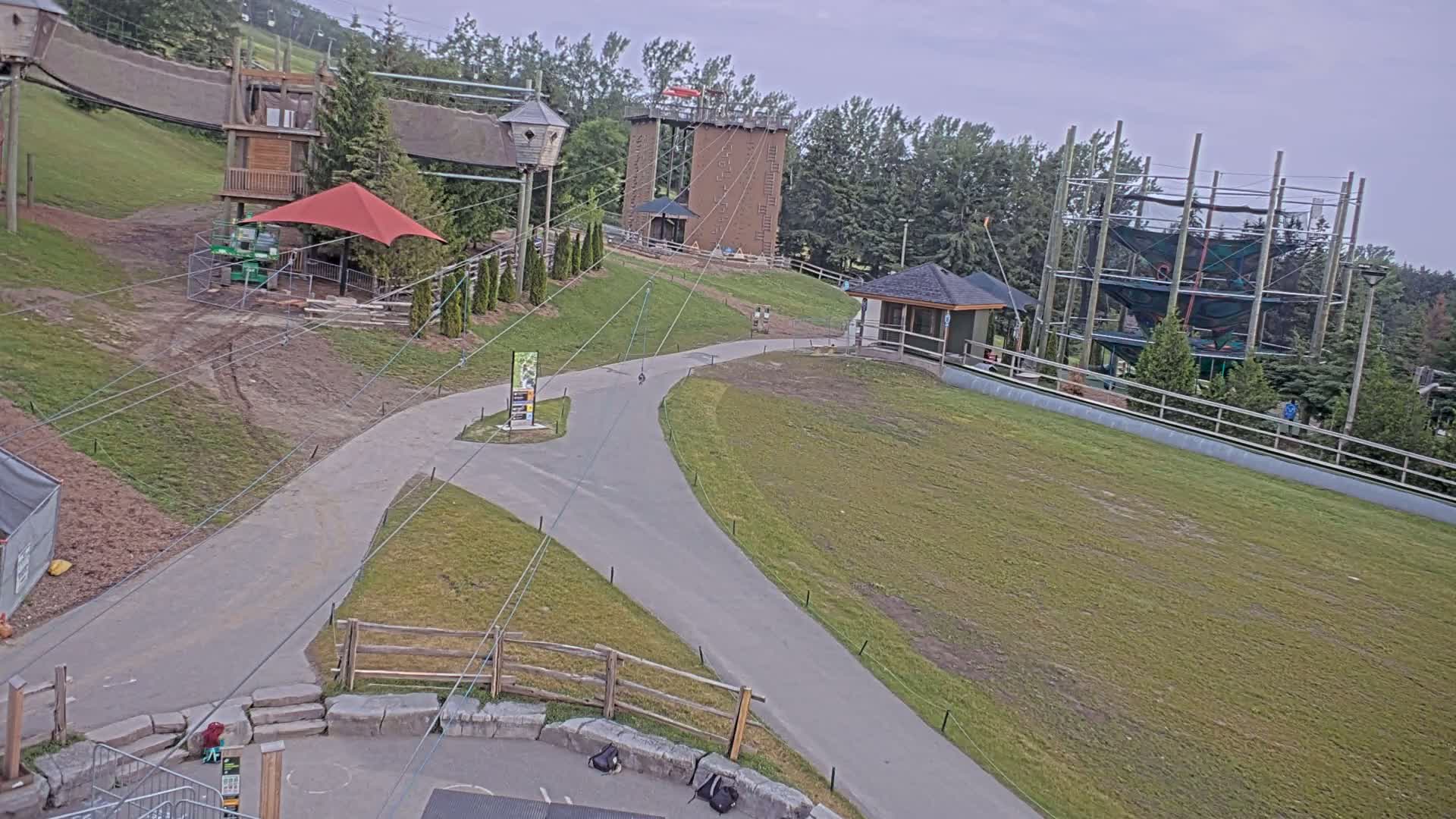 An outdoor aerial view shows a paved pathway winding through a grassy area with various play structures, including climbing walls and a ropes course, under an overcast sky.